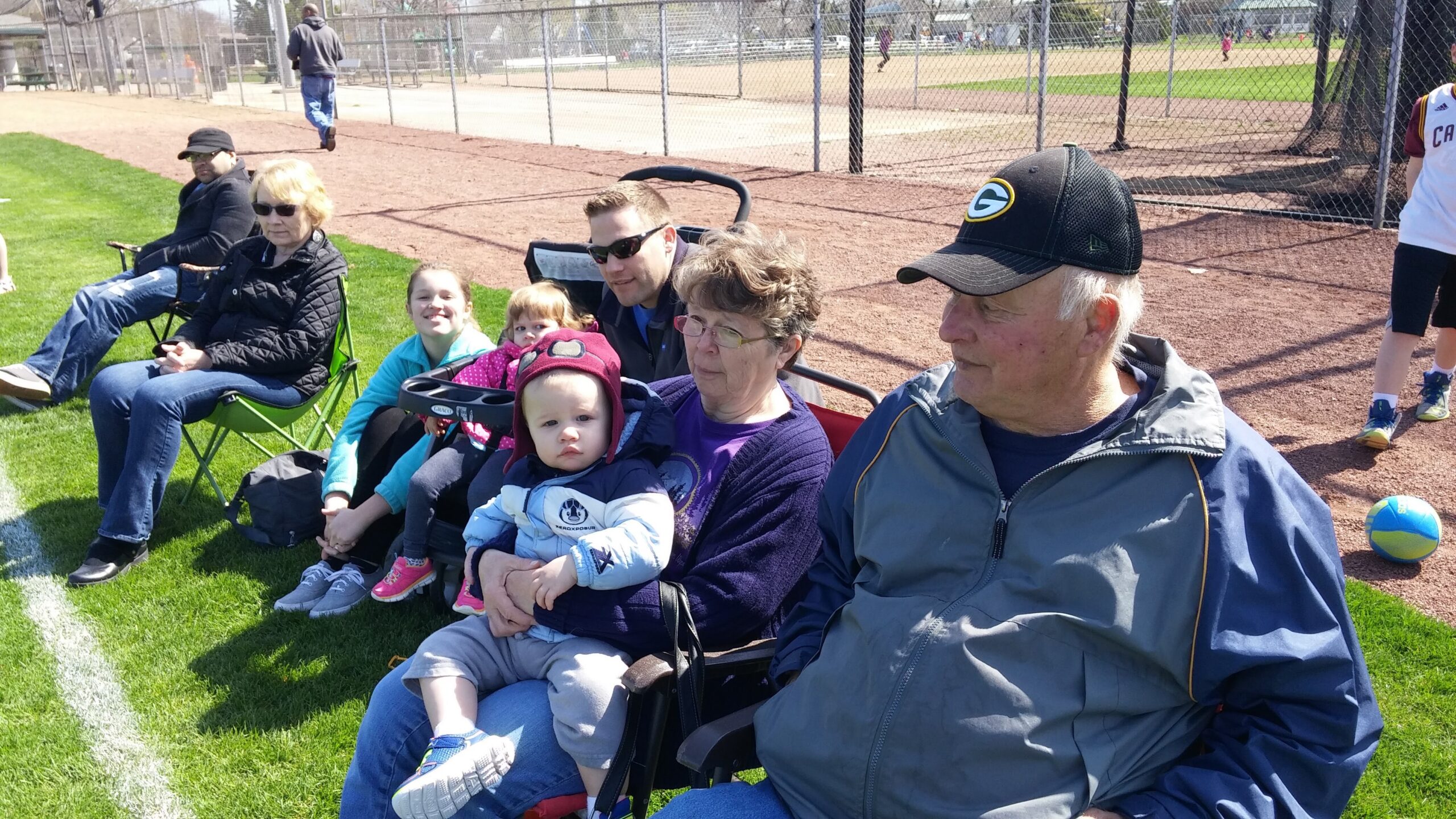 Group of people seated on foldable chairs and a stroller on a grassy field, watching a baseball game behind a chain-link fence.