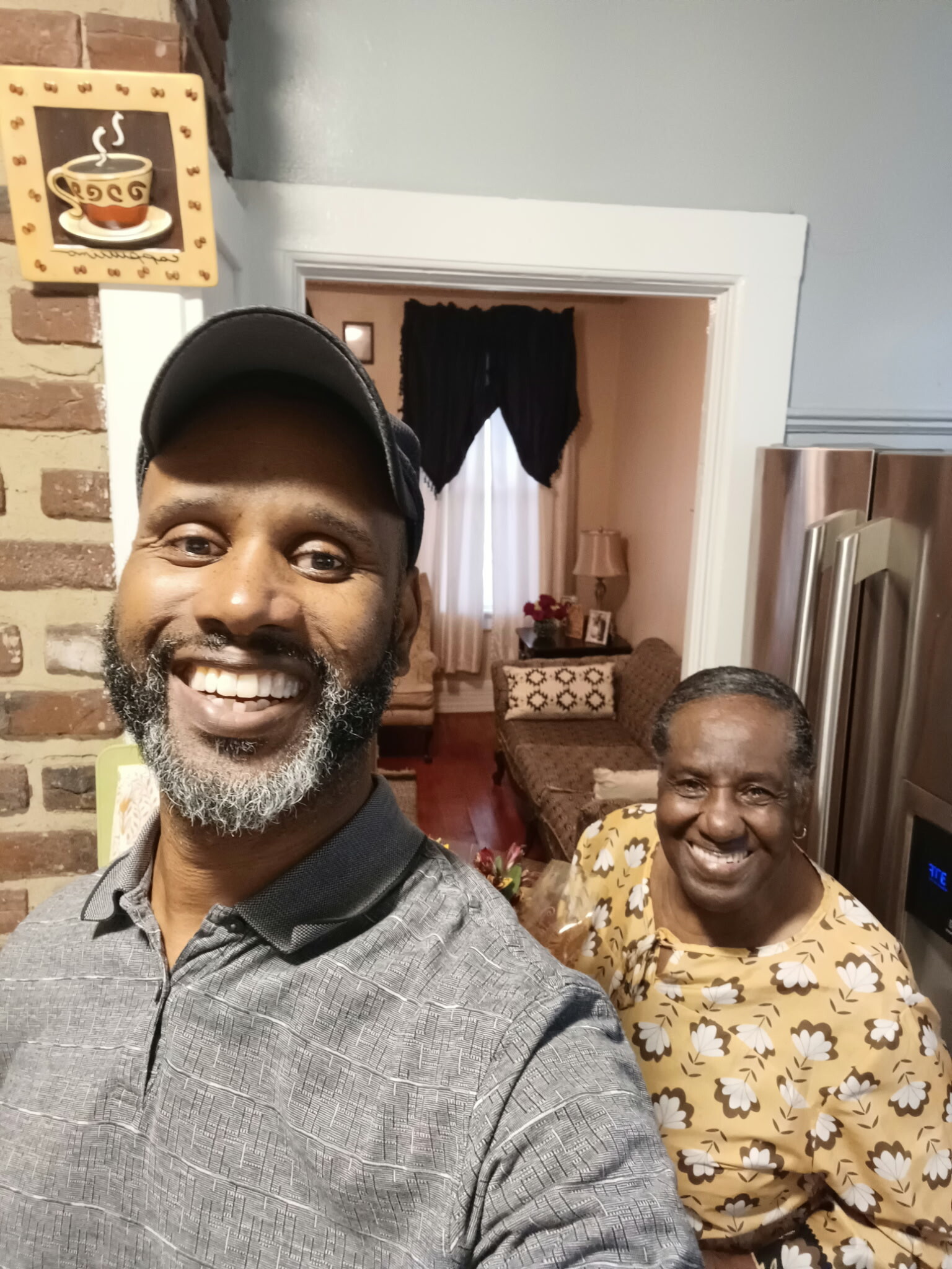 Selfie of a smiling man in a gray shirt and cap in the foreground, with a smiling woman in a yellow floral dress in the living room behind him by a doorway to a cozy family room.