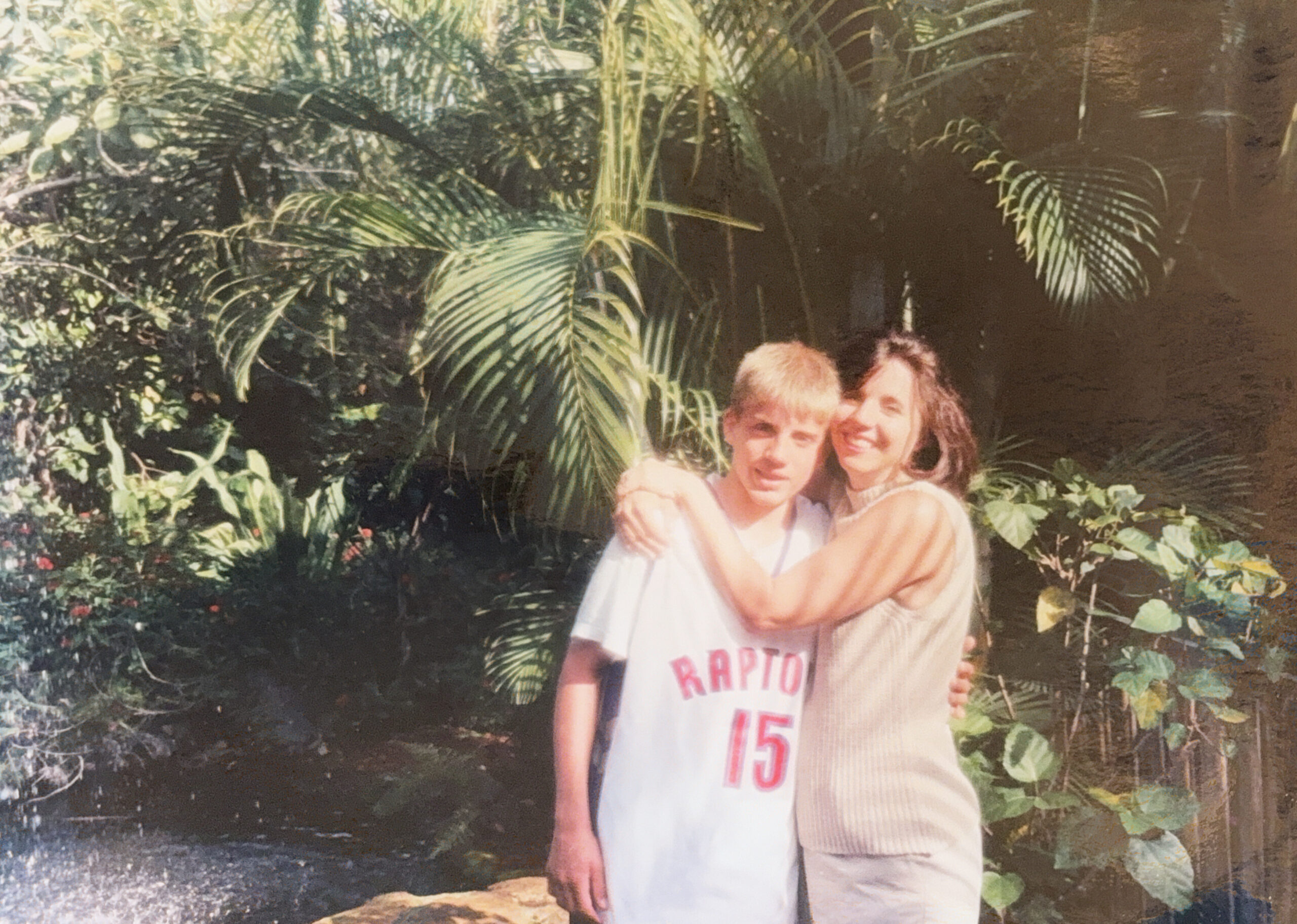 Two friends posing outdoors in a tropical garden, one wearing a white Raptors basketball jersey with the number 15, smiling at the camera.