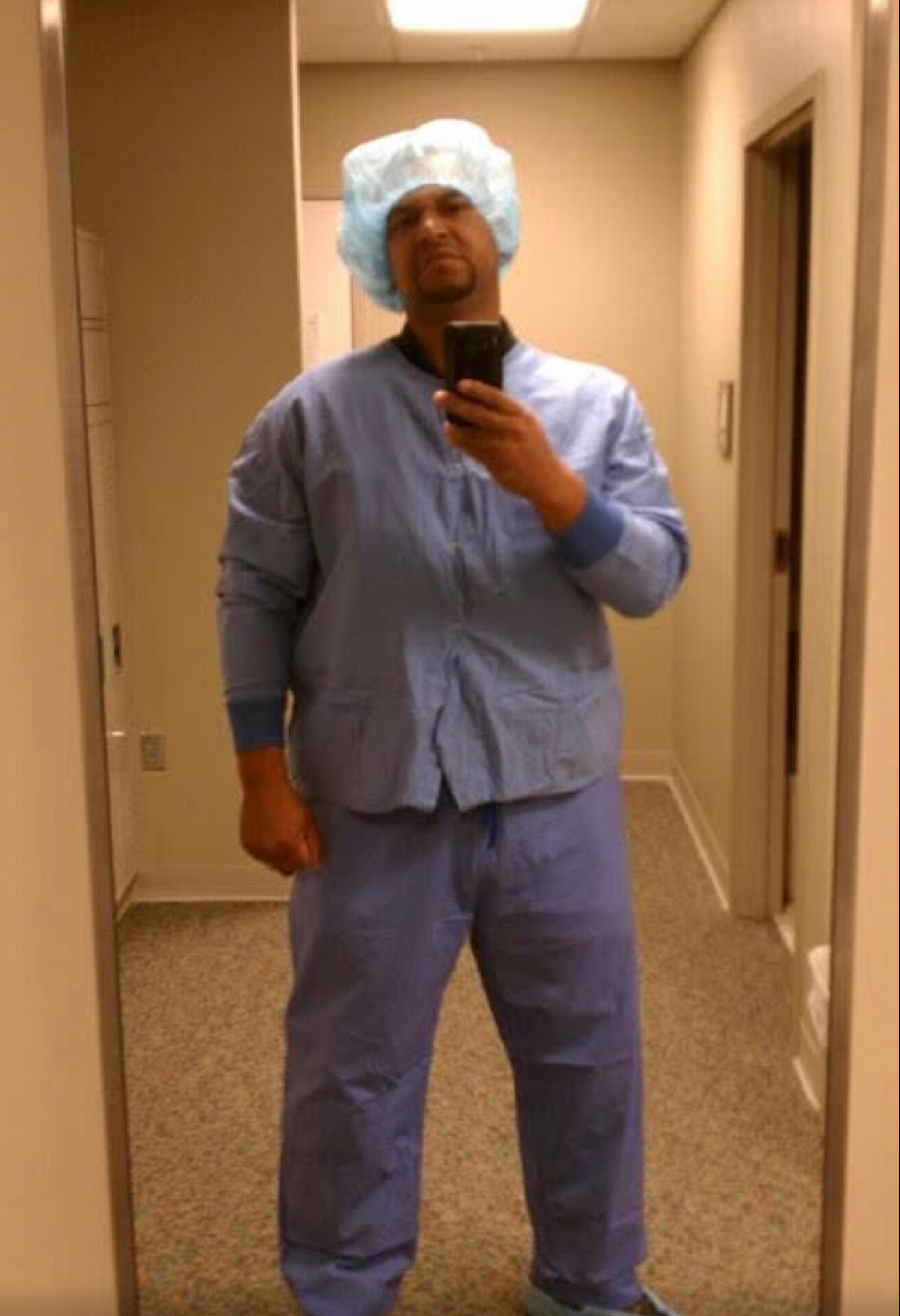 Healthcare worker in blue scrubs and a disposable hairnet taking a mirror selfie in a hallway, holding a smartphone.