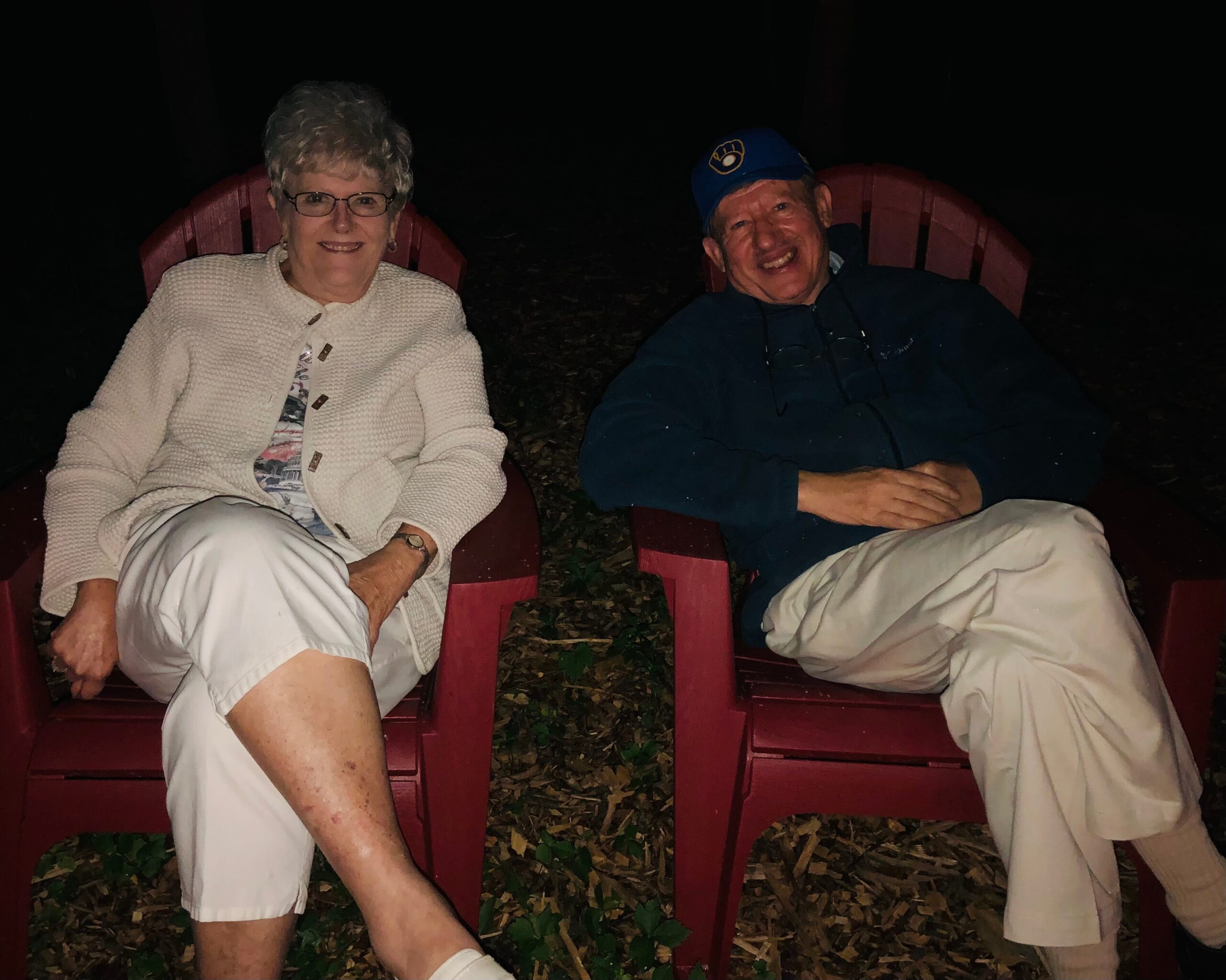 Two elderly adults sitting in red outdoor chairs at night, smiling at the camera likely enjoying a chilly evening outdoors.