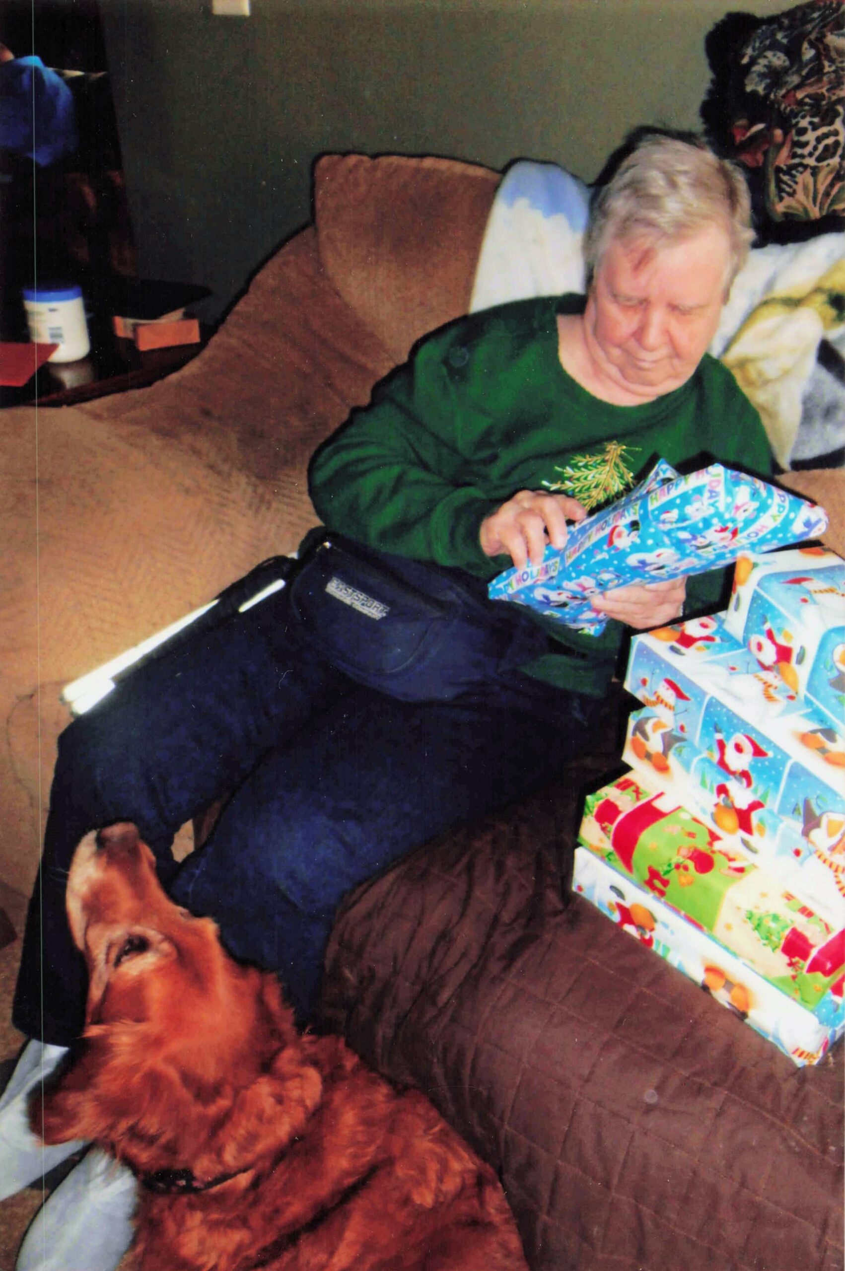 Older person in a green sweater sits on a brown couch, unwrapping a blue gift while a brown dog lies nearby on the floor with wrapped presents nearby.
