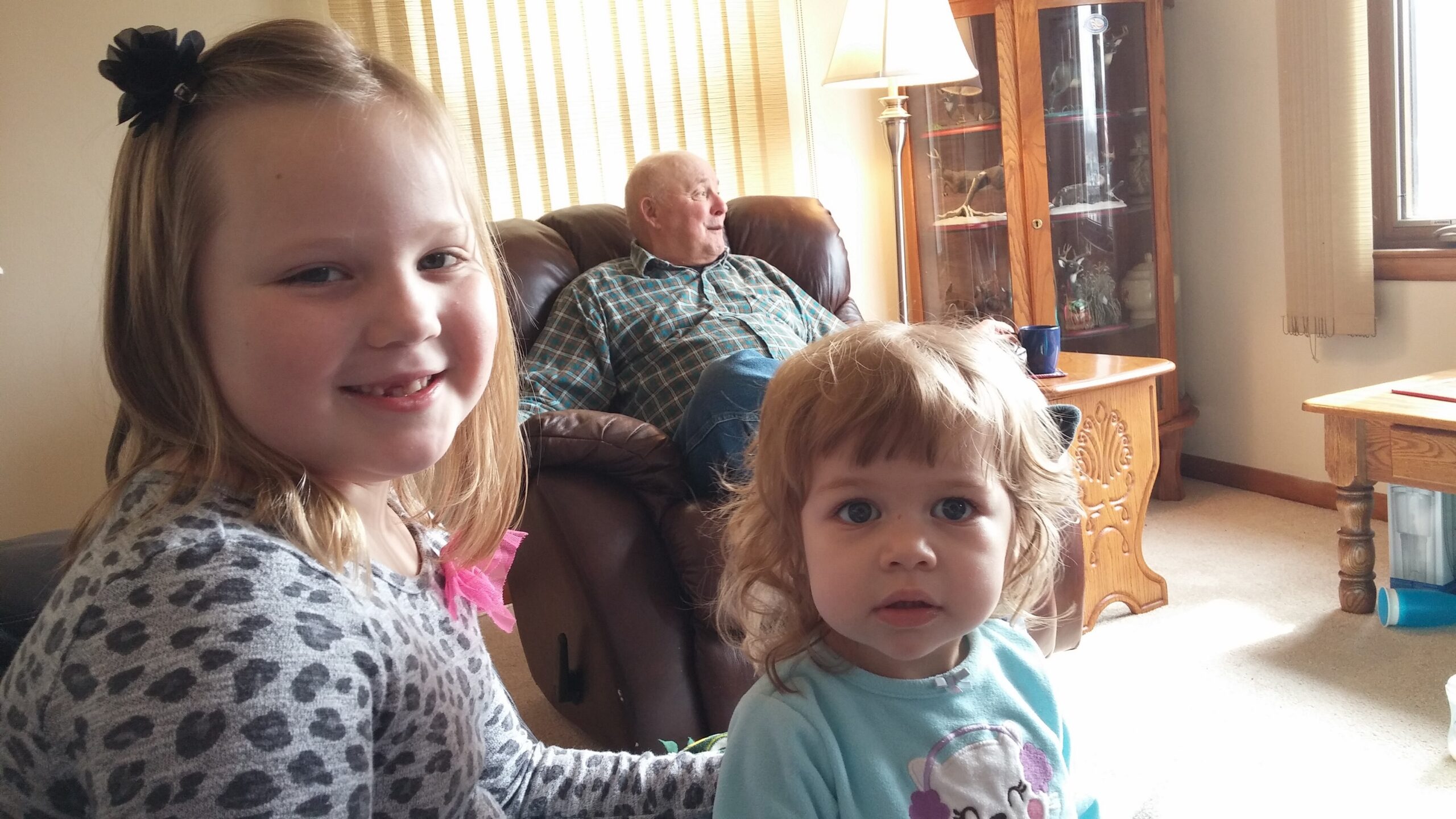 Three young children pose for a selfie in a living room with an older man seated on a brown leather chair in the background.