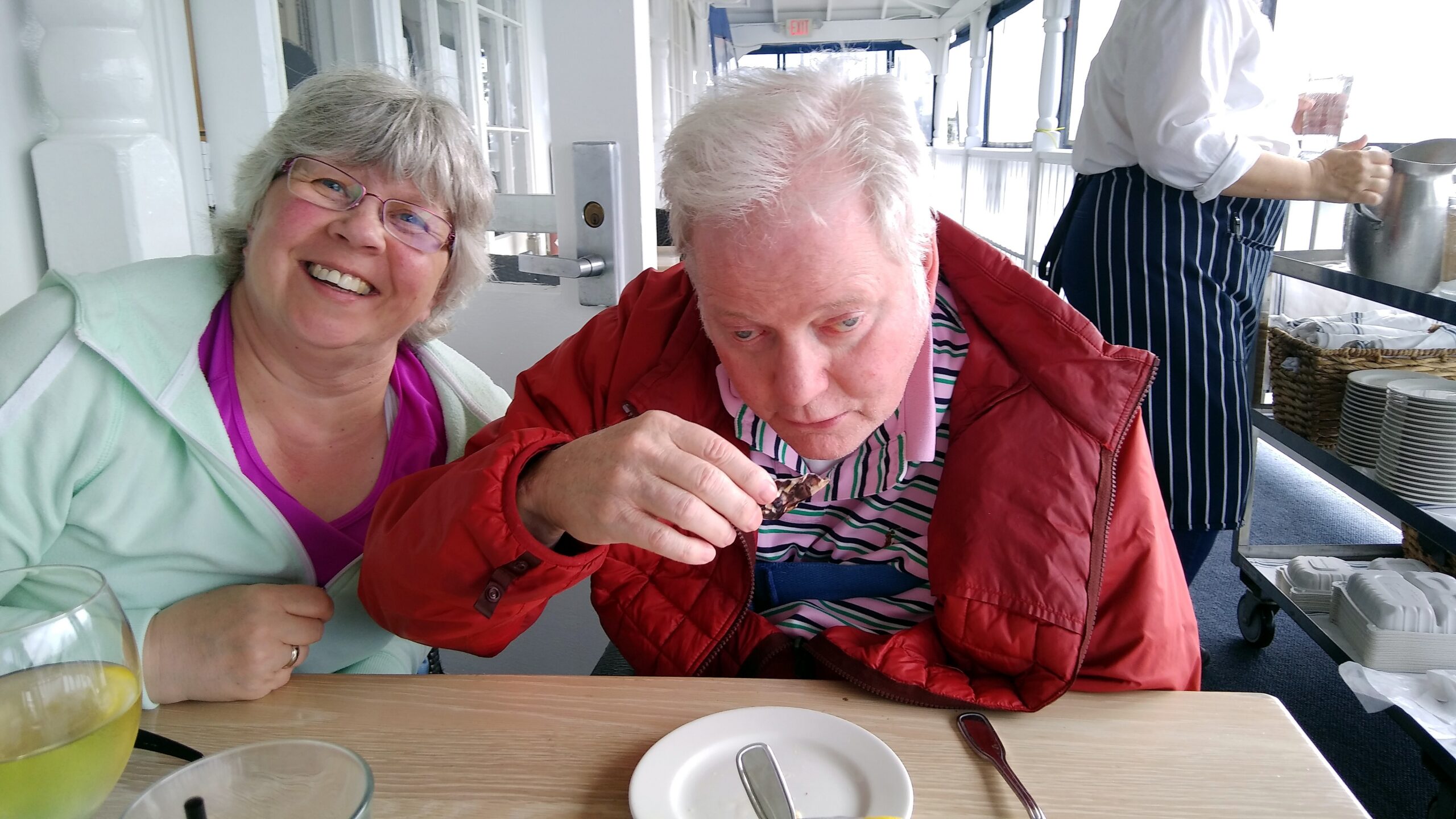 Smiling woman with pink glasses sits beside an older man in a red jacket as they share dessert on a boat dining deck.