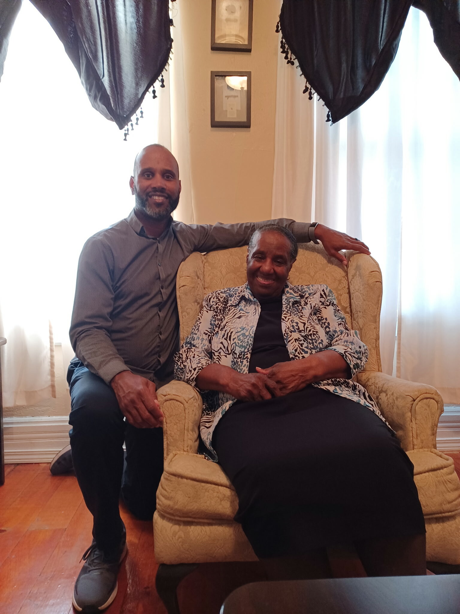 Smiling man kneels beside an elderly woman sitting in a beige patterned armchair in a sunlit living room.