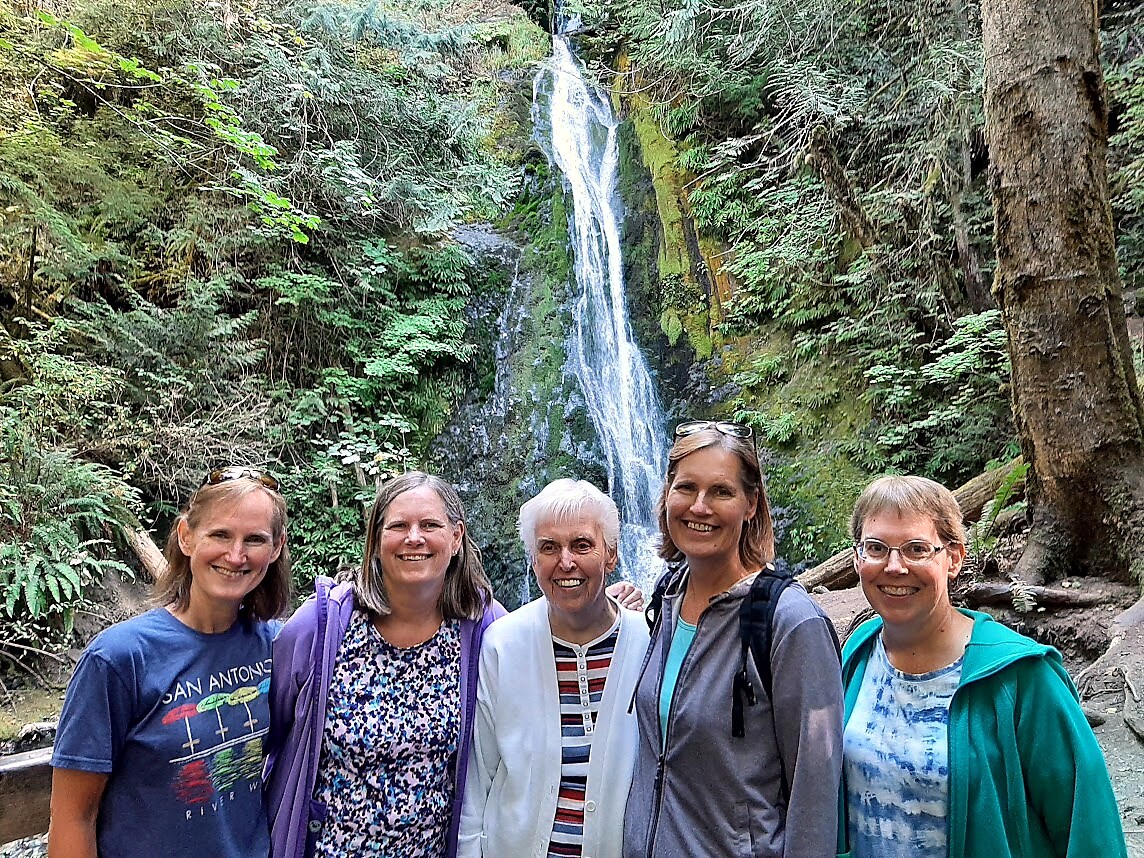 Five women stand together in a forest, smiling, with a tall waterfall behind them.