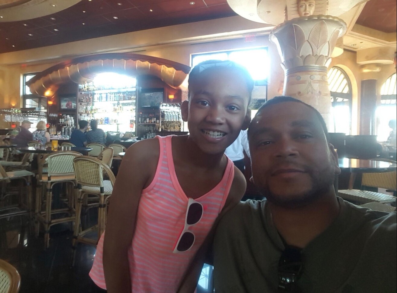 Two people pose for a selfie in a tropical restaurant/bar with wicker chairs and a bar in the background.