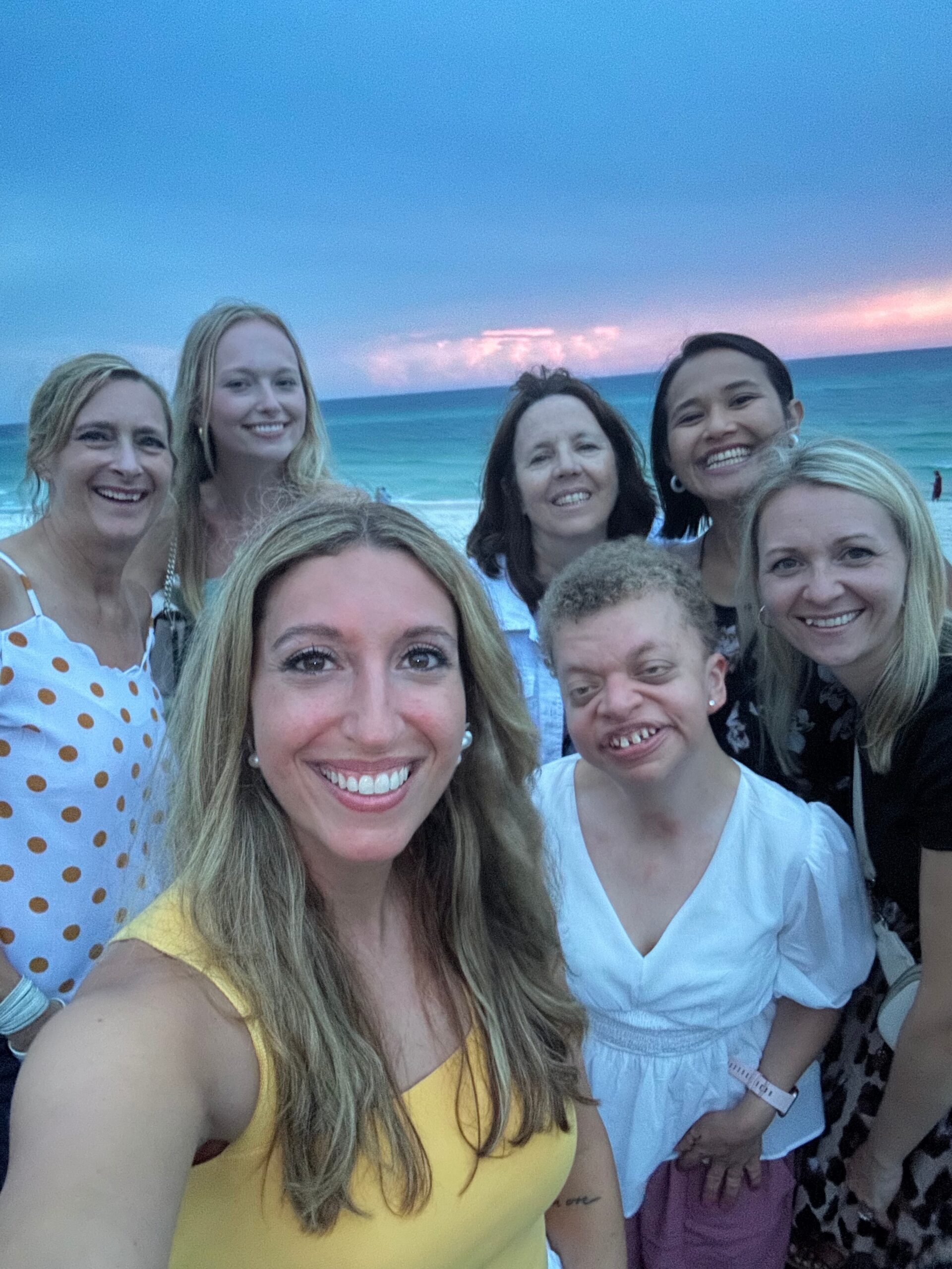 Group of smiling women taking a selfie on a sandy beach with the ocean and a pink sunset in the background.