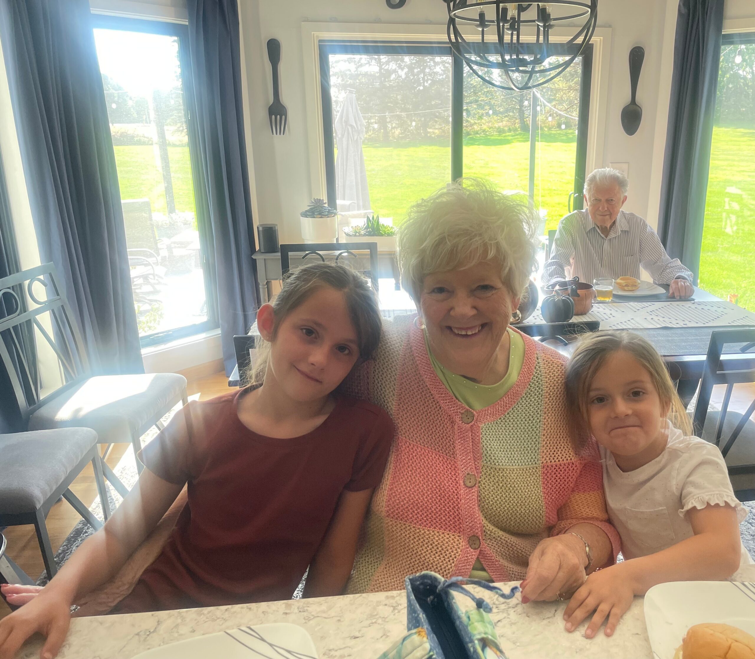 Smiling family of four seated at a marble table indoors, sunlit windows and a green yard outside behind them, with pastries on the table.