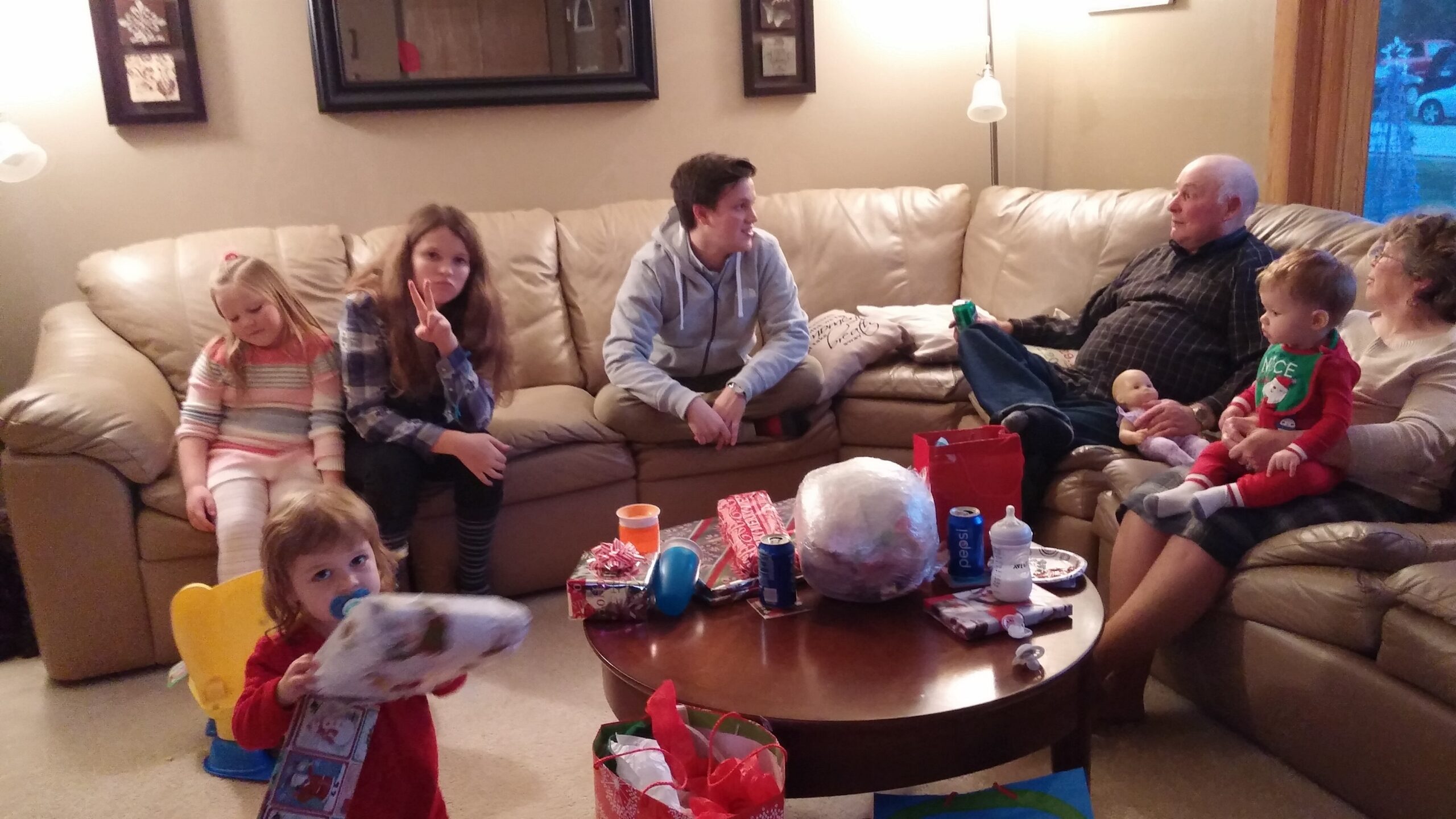 Family scene in living room with kids and adults on a beige sectional, opening gifts and chatting during a cozy gathering.