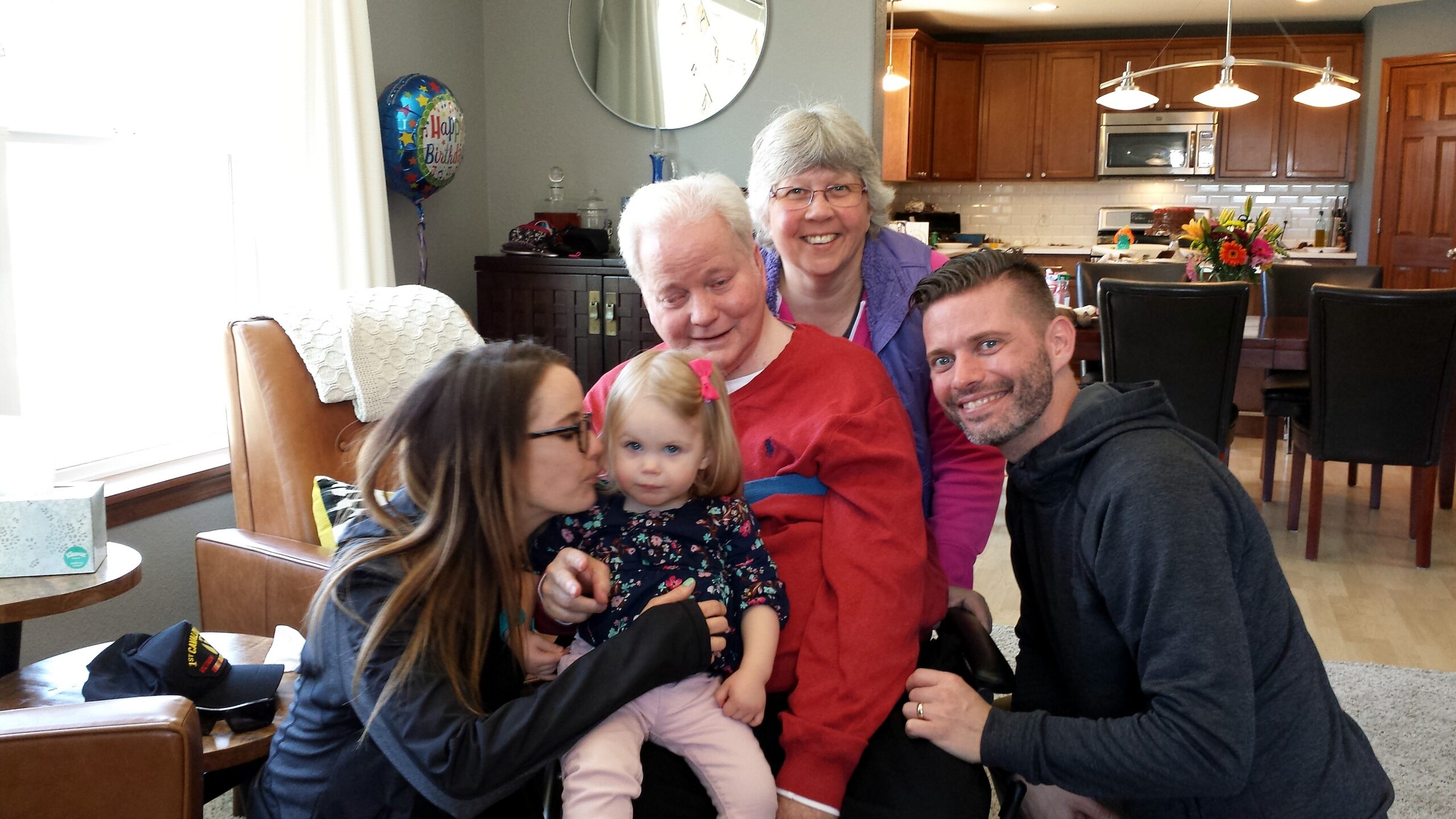 Smiling family posing together in a kitchen-dining area, with a birthday balloon in the background and a young girl sitting on a woman's lap.
