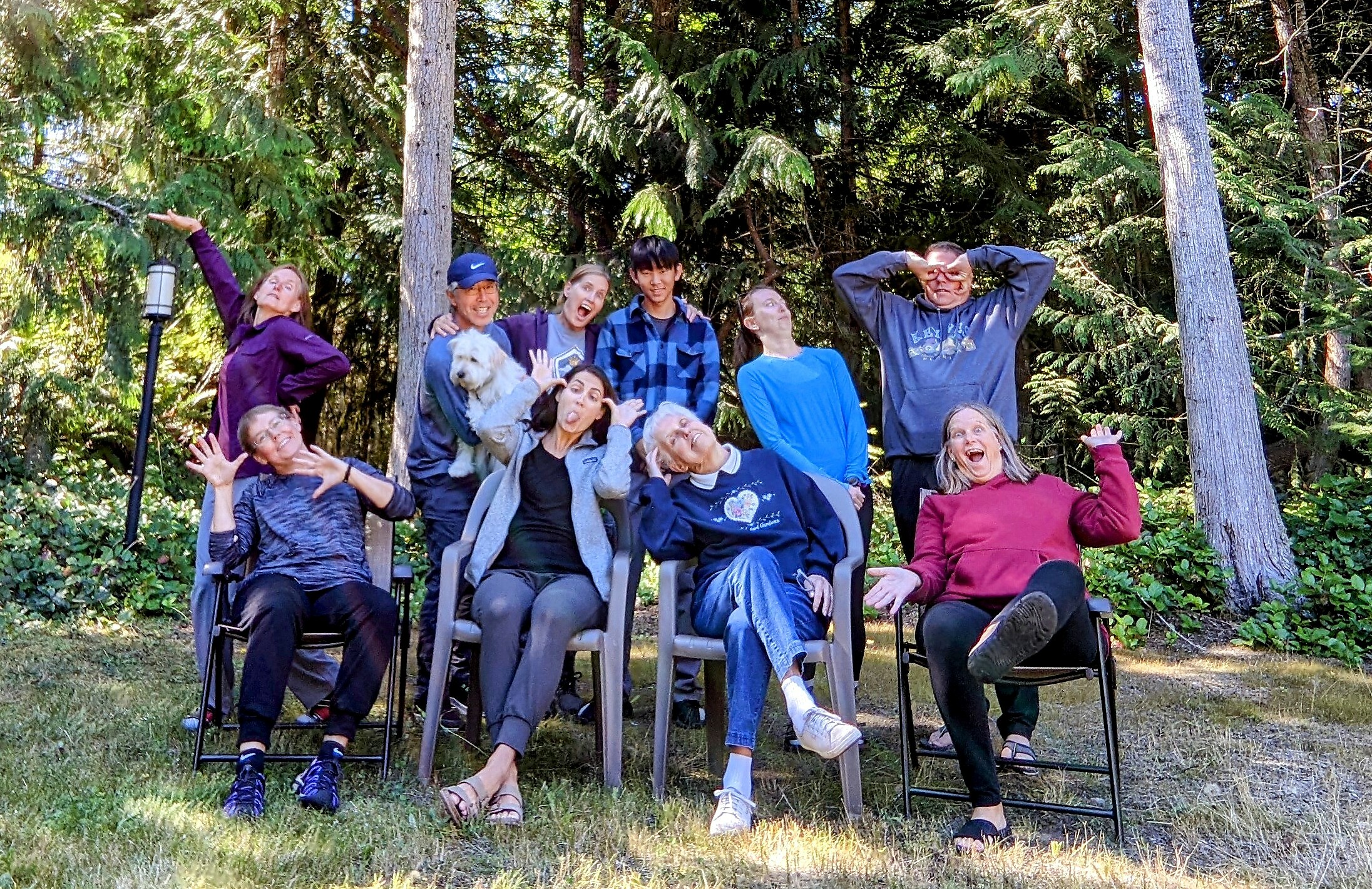 Group of adults posing in a wooded backyard, seated in lawn chairs and making goofy gestures.