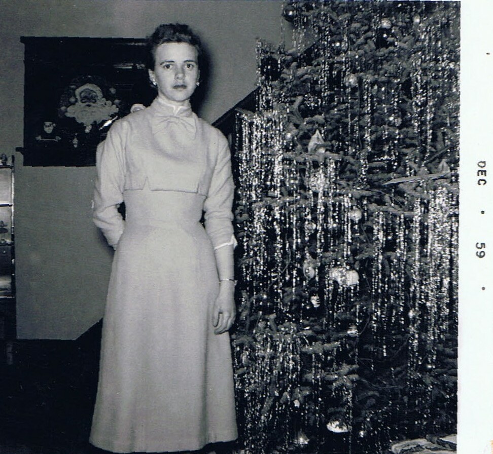 Woman in a long light dress standing beside a Christmas tree decorated with tinsel in a living room (black-and-white photo).