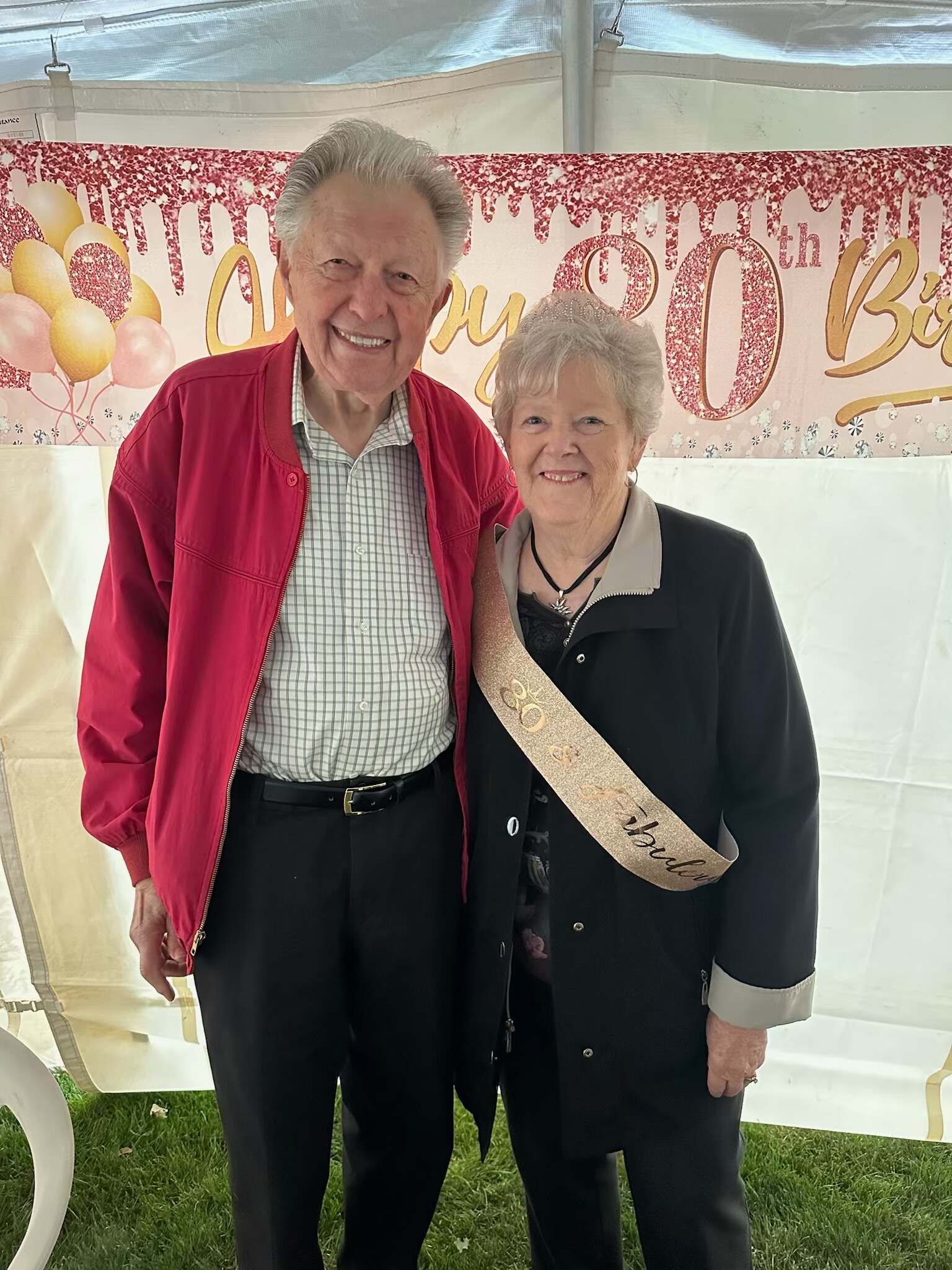 Two smiling seniors at a 60th birthday party; man in a red jacket and checkered shirt, woman wearing a tiara and a sash.