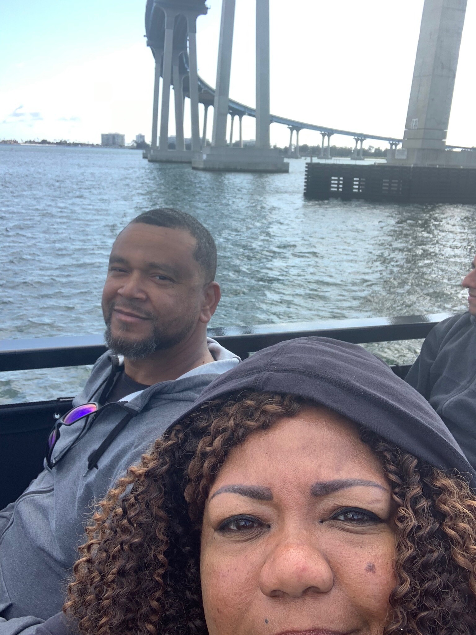 Couple on a boat, man smiling in the background and woman in the foreground, with a large bridge over water.