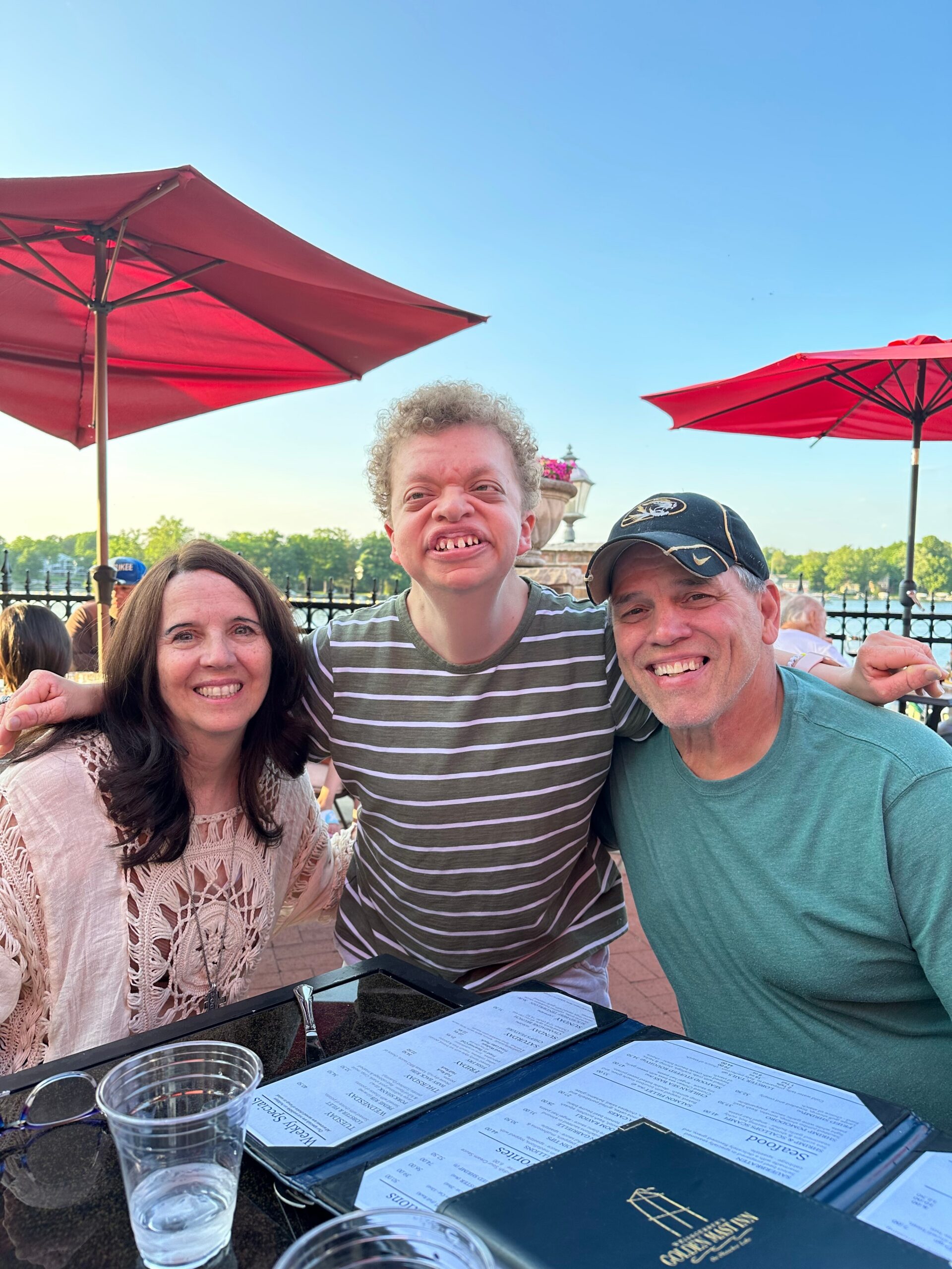 Three people smile for the camera at an outdoor cafe with red patio umbrellas and a menu on the table in front of them.