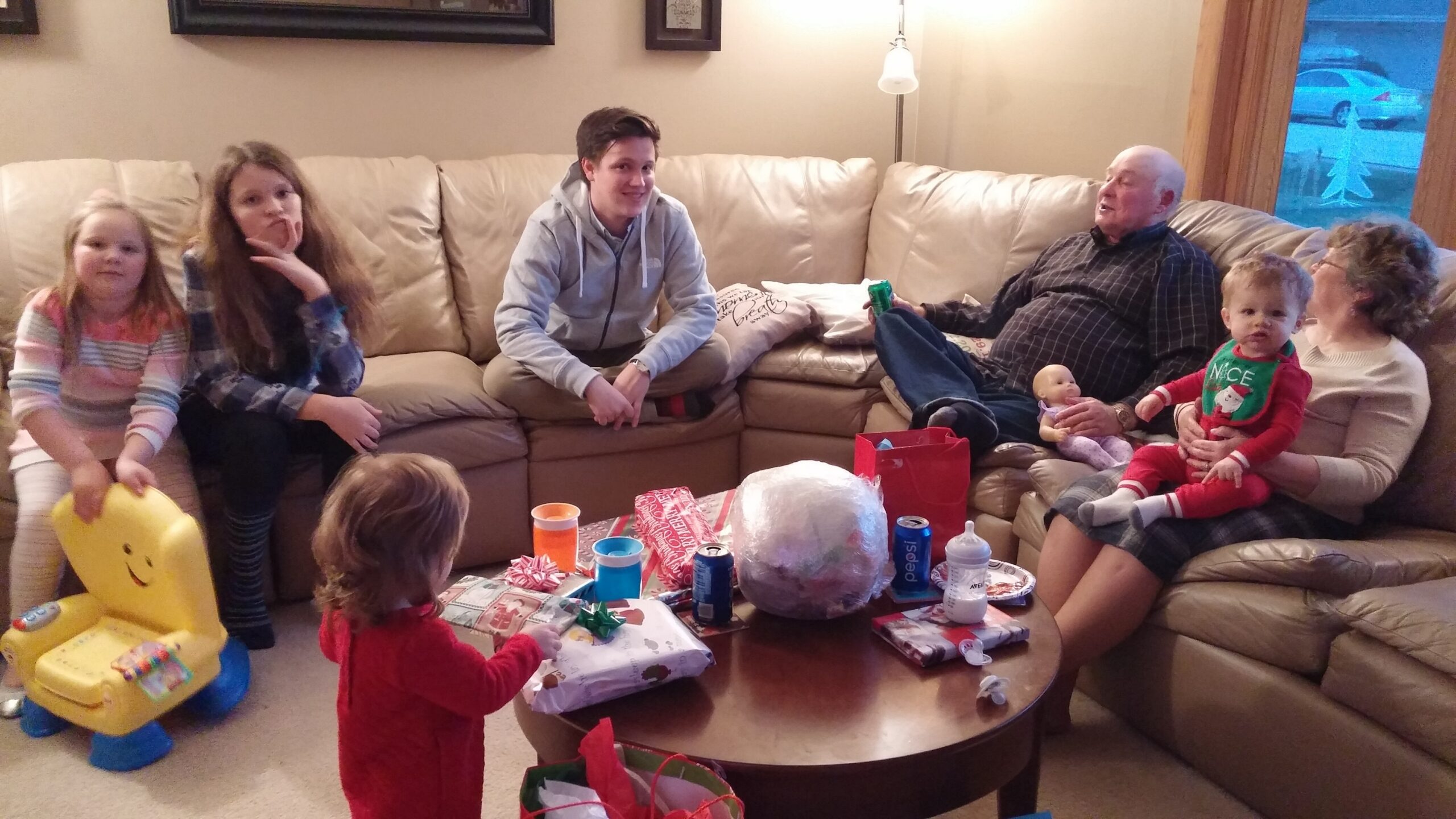 Family gathered in living room for a gift exchange; kids on a tan sofa, adults nearby, table full of presents and snacks.