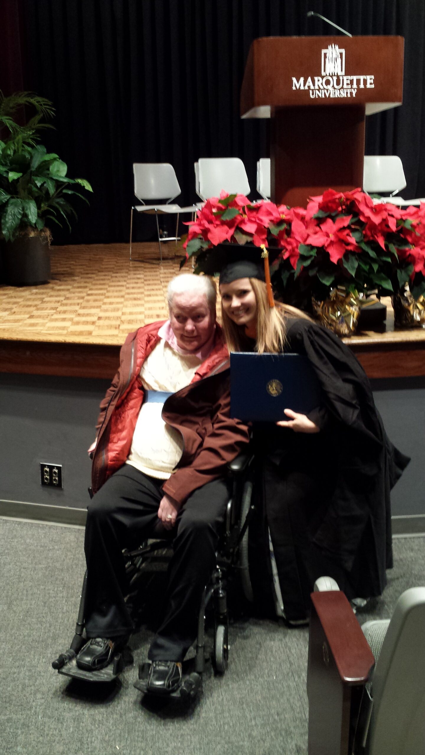 Graduate in cap and gown posing with an elderly man in a wheelchair on a stage with a Marquette University podium and red poinsettias in the background