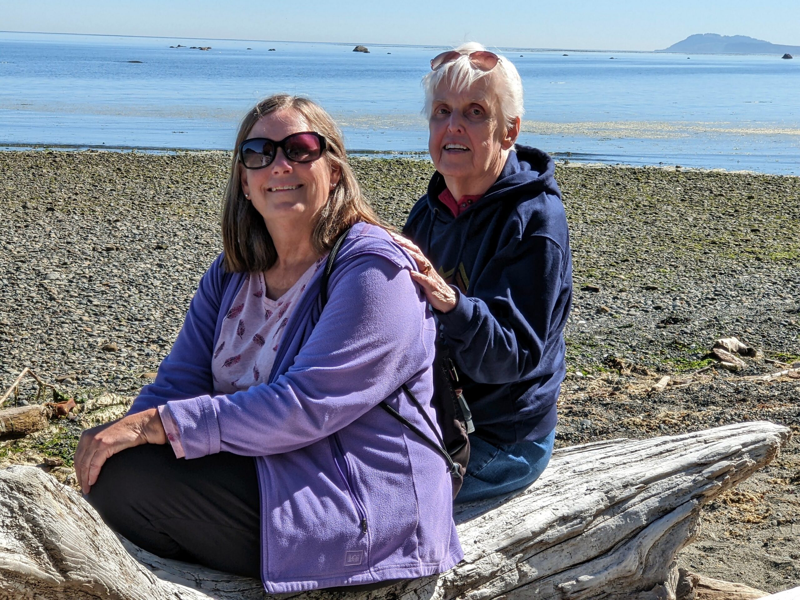 Two smiling women pose on a weathered driftwood log at a rocky beach, with calm blue water and a distant island on the horizon.