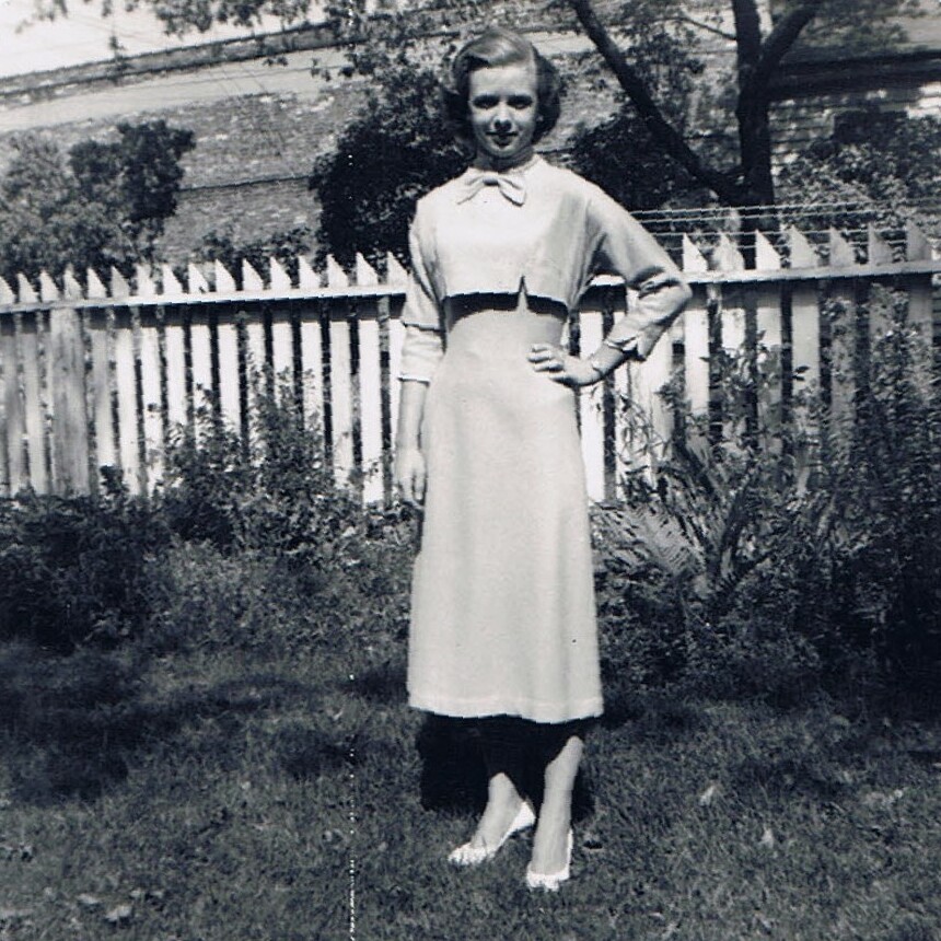 Woman in a vintage knee-length dress with a bow at the neckline, standing on grass in front of a white picket fence.