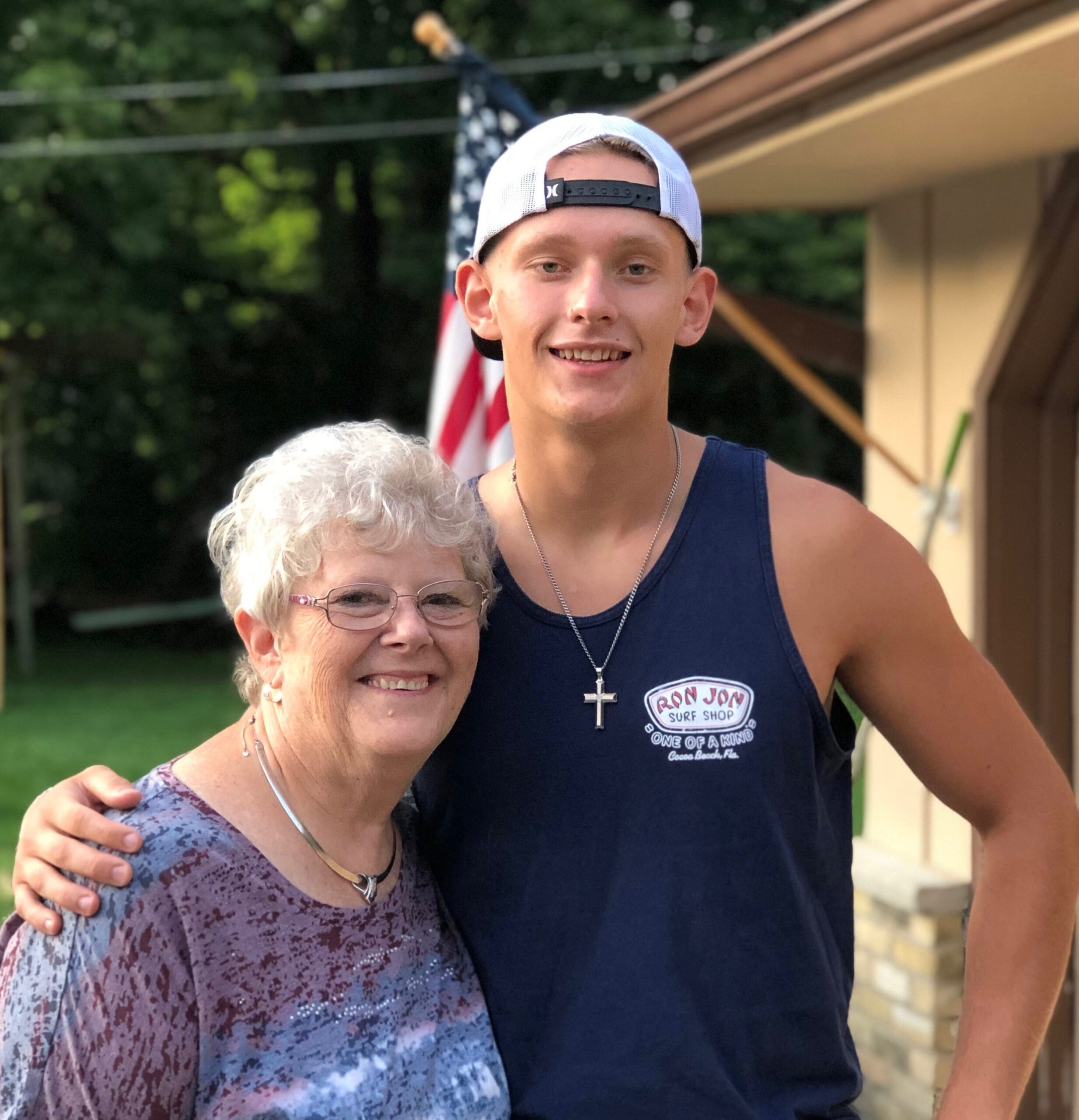 Two people smiling outdoors: an older woman in a purple patterned top in the foreground and a young man in a navy tank top and white cap behind her, with an American flag in the background.