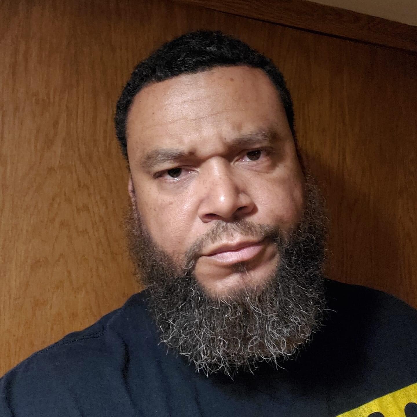 Close-up of a man with a full beard and mustache looking slightly to the left, neutral expression, dark shirt, wooden background.
