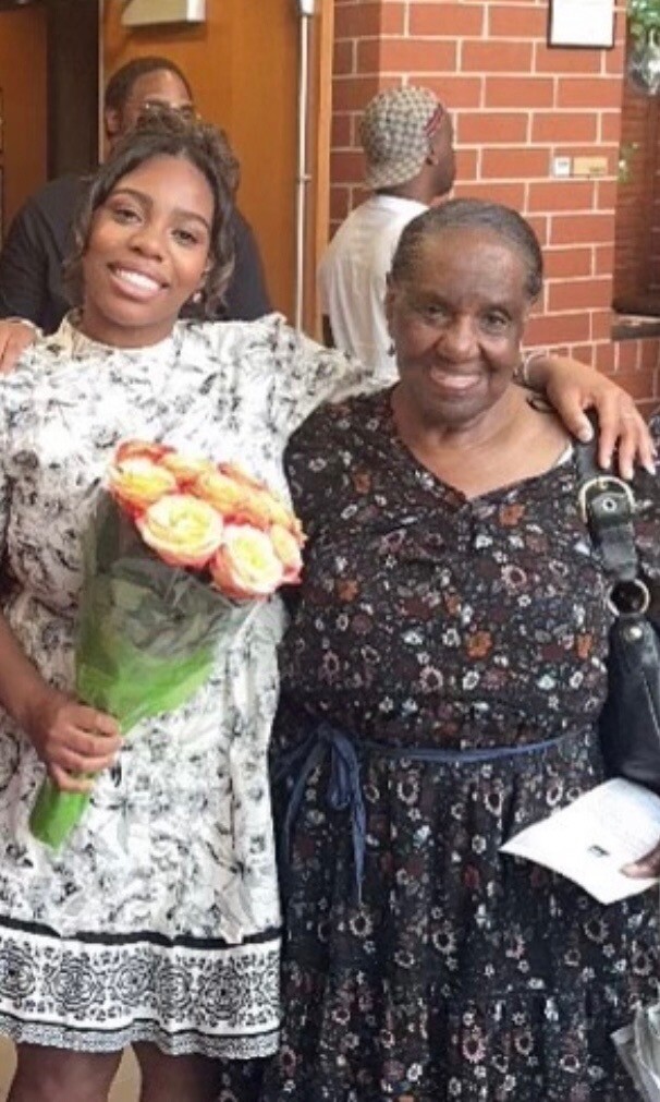 Two women smile and pose with their arms around each other; the younger woman holds a bouquet of peach and pink roses indoors.