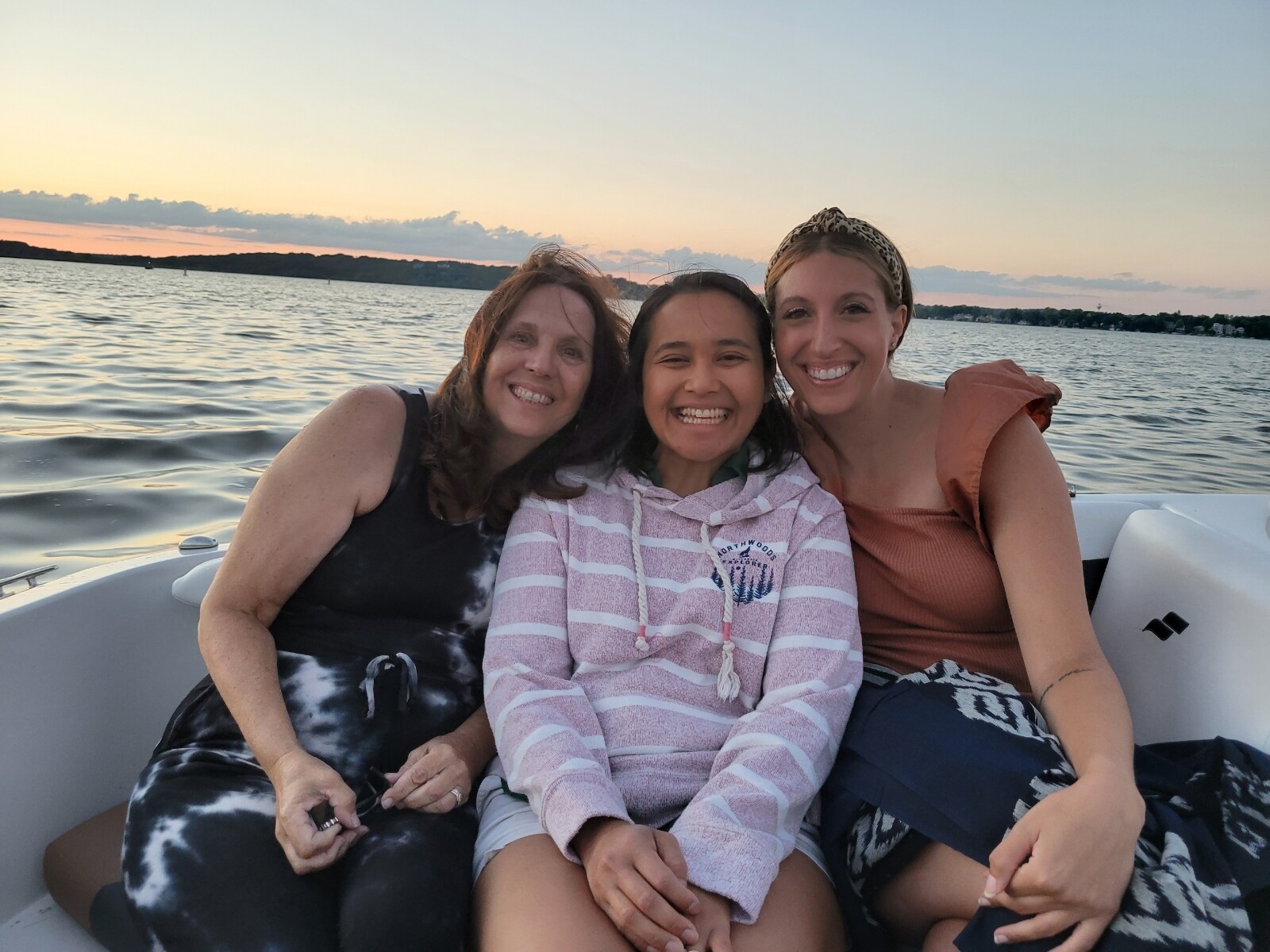 Three smiling women sitting on a boat at sunset with water and a distant shore in the background.