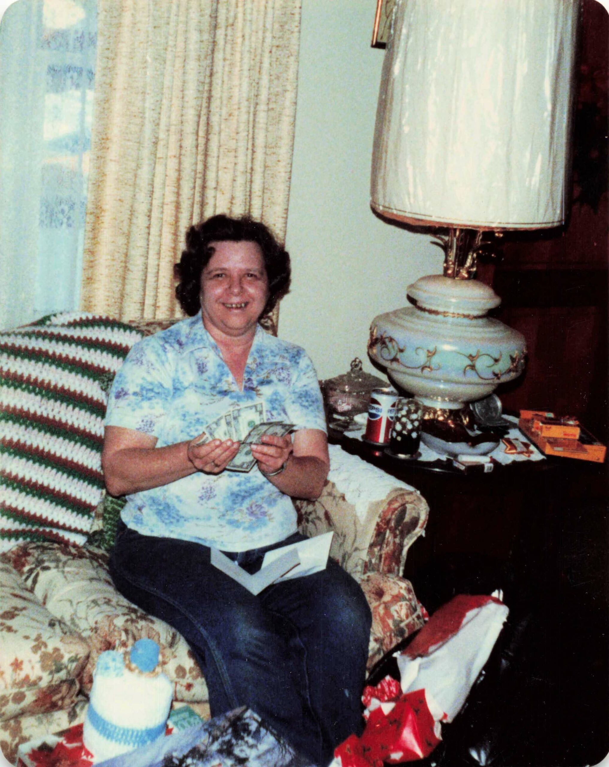 Smiling woman in a blue floral shirt sitting on a patterned sofa, counting money beside a large decorative lamp and wrapped gifts nearby.