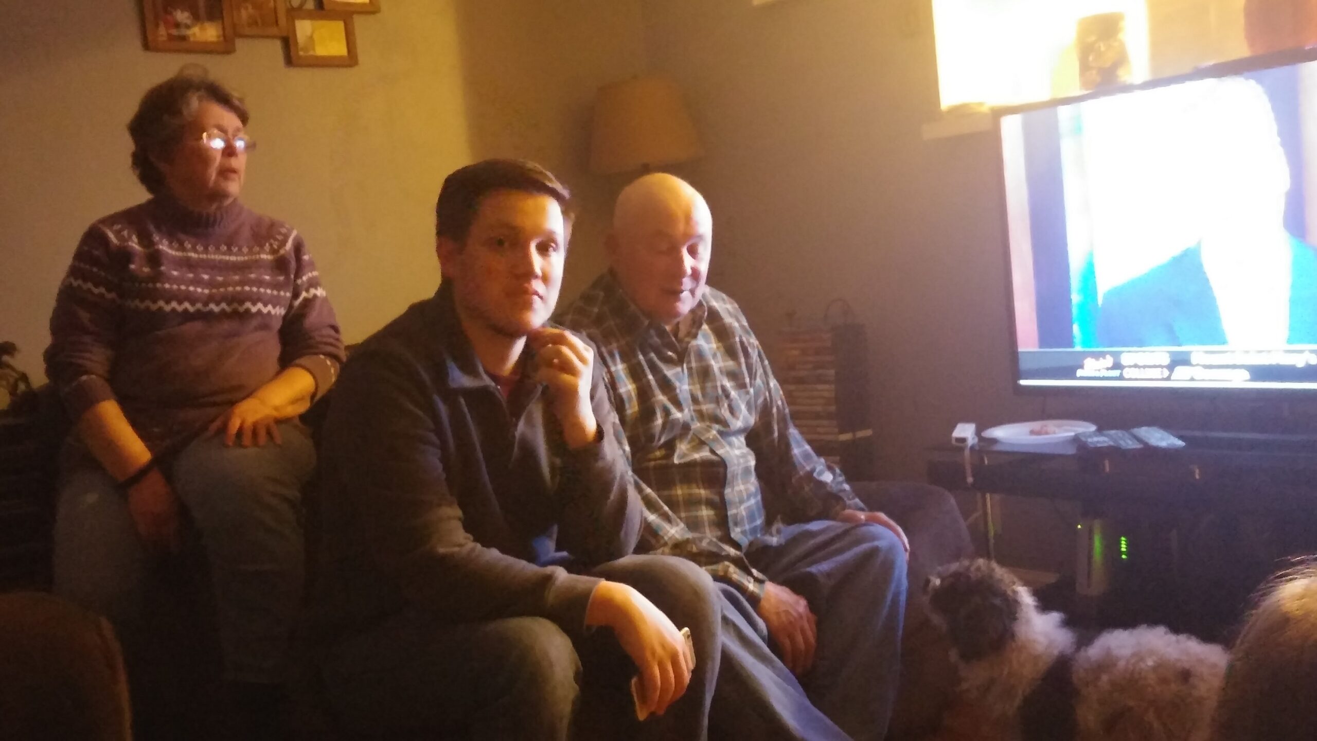 Three adults sit on a couch in a dim living room watching a large TV screen, warm lamps behind them.