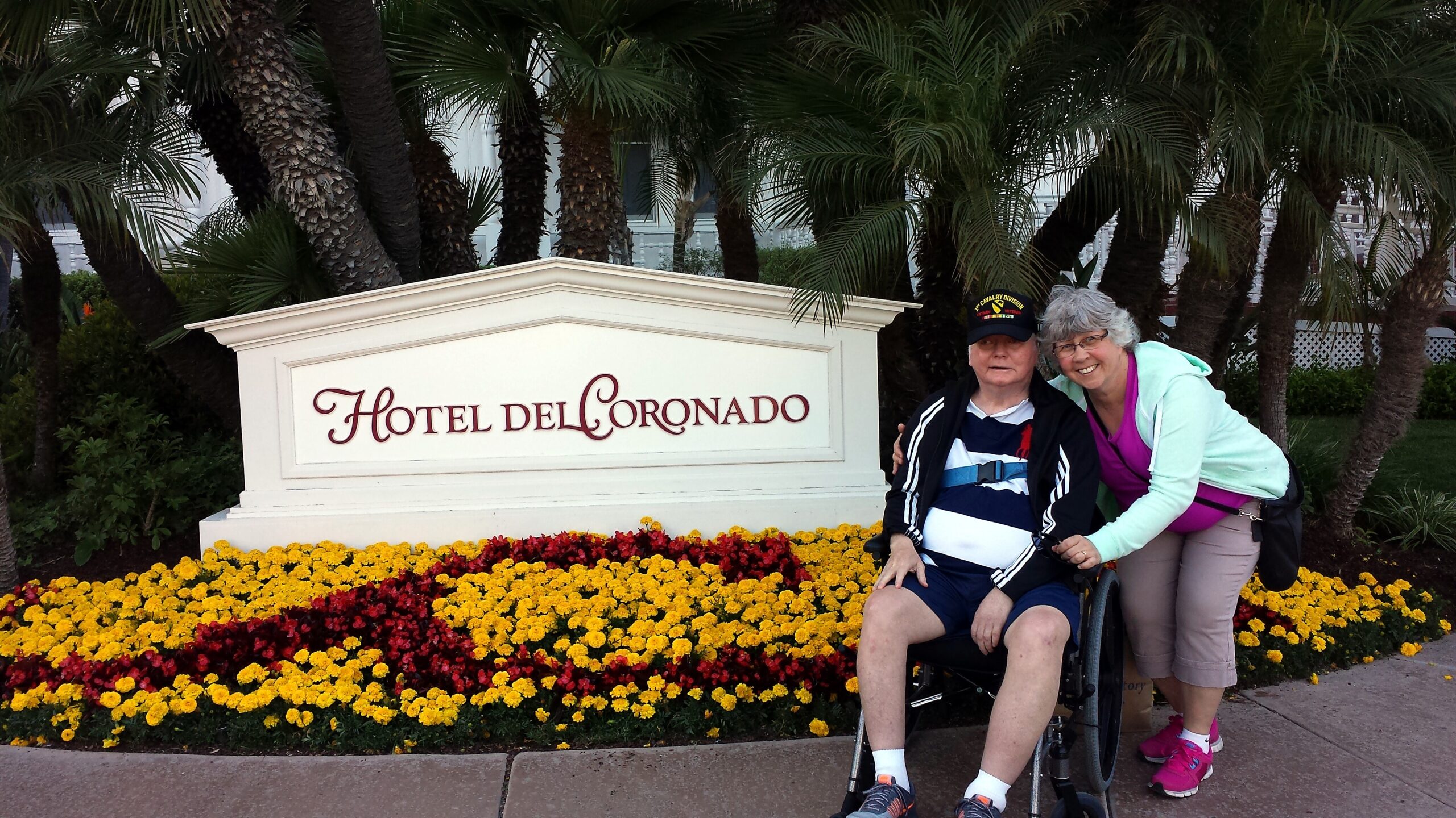Two visitors, one in a wheelchair, pose beside a white sign that reads 'Hotel del Coronado' amid palm trees and a bed of yellow and red flowers.