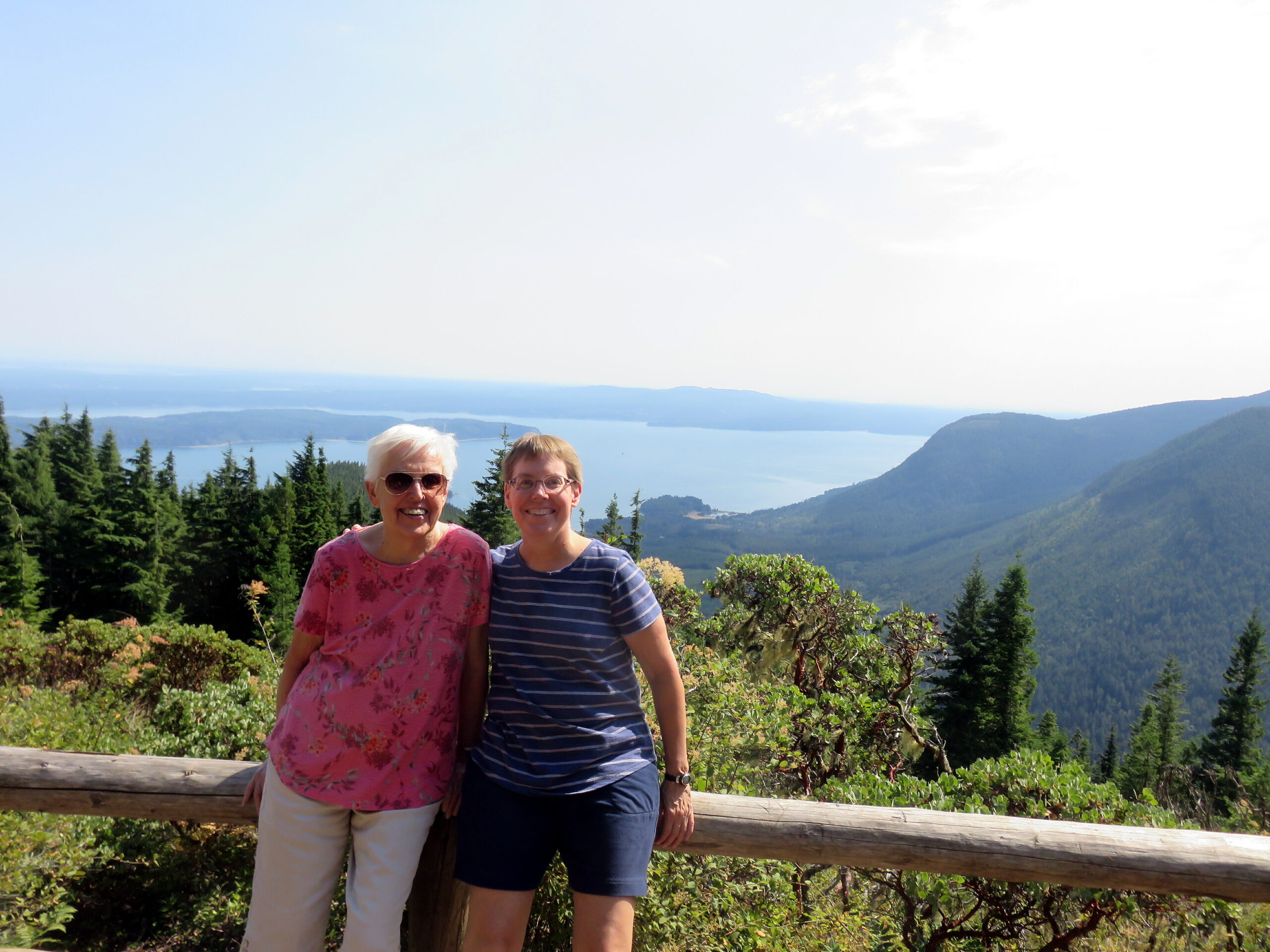 Two women posing together outdoors on a hillside with trees, a wooden railing, and a lake and mountains in the distance.