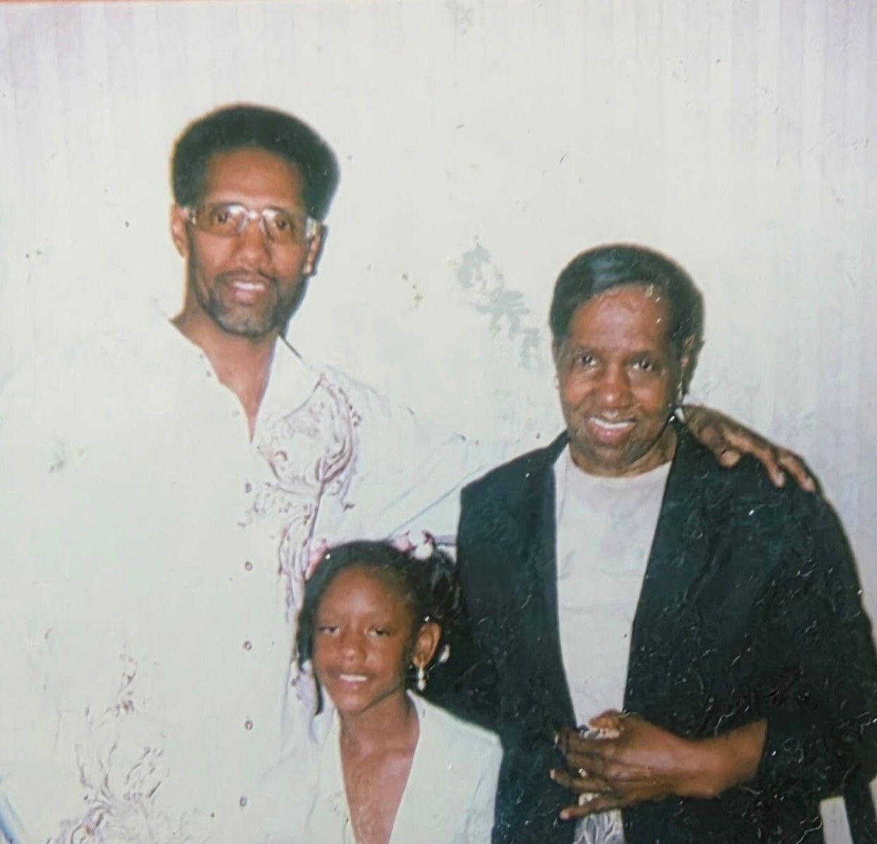 Family portrait of three people posing together: a man with glasses on the left, a smiling man in a dark blazer on the right, and a young girl in front.