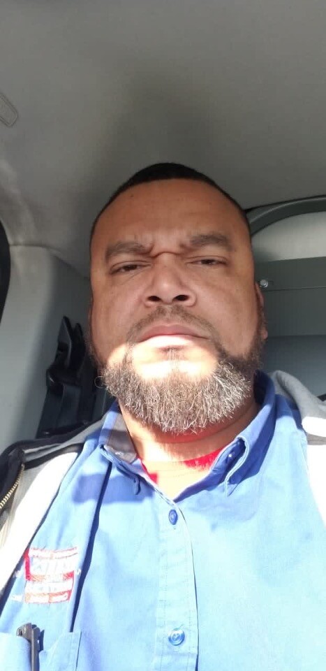 Close-up of a man with a gray beard in a blue collared shirt inside a vehicle, looking at the camera with a serious expression.