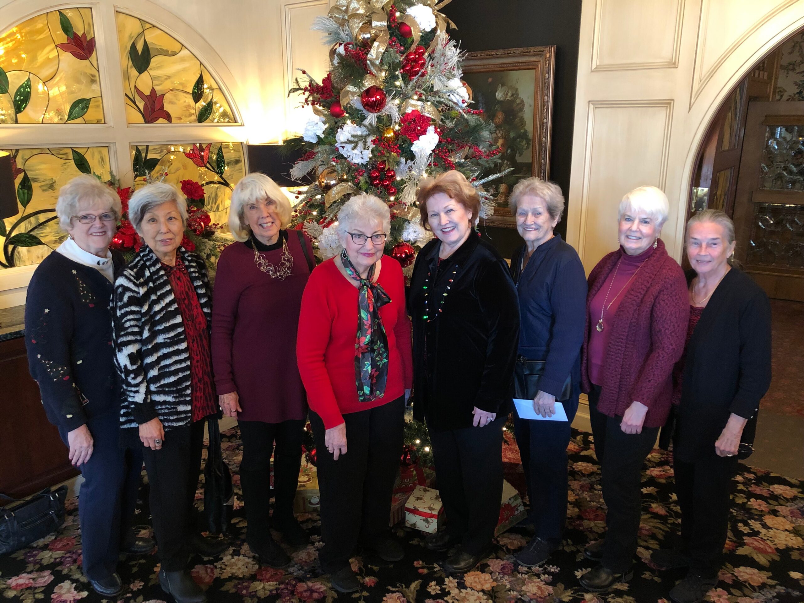 Group of eight women posing for a holiday photo in front of a decorated Christmas tree with red ornaments, ribbons, and presents underneath in an elegant lobby.