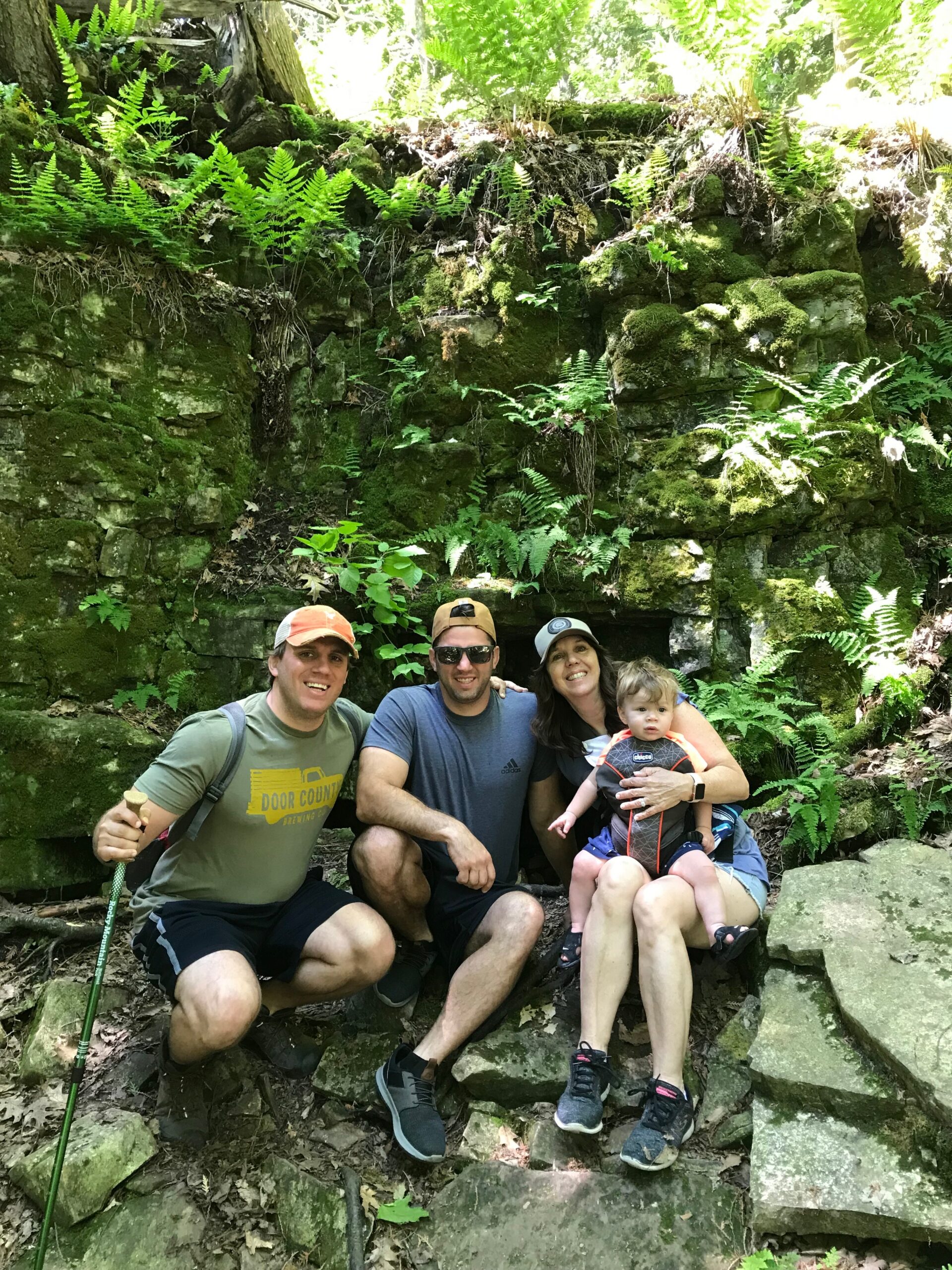 Family of five posing on mossy rock wall in a lush, fern-filled forest; adults with a baby in a carrier are smiling at the camera while outdoor gear is visible nearby.