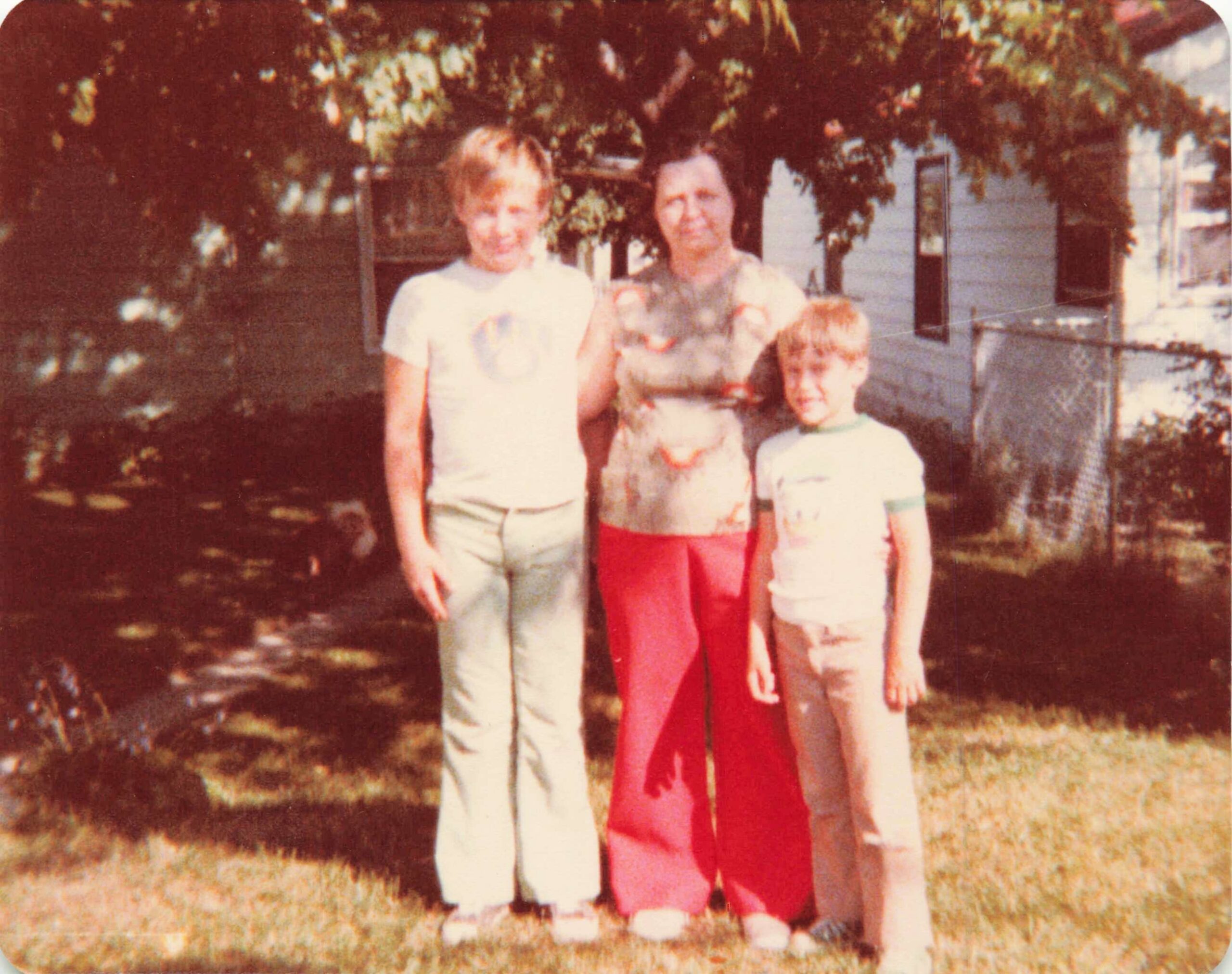 Three people—a woman and two children—pose outside in a yard with a tree and a house in the background.
