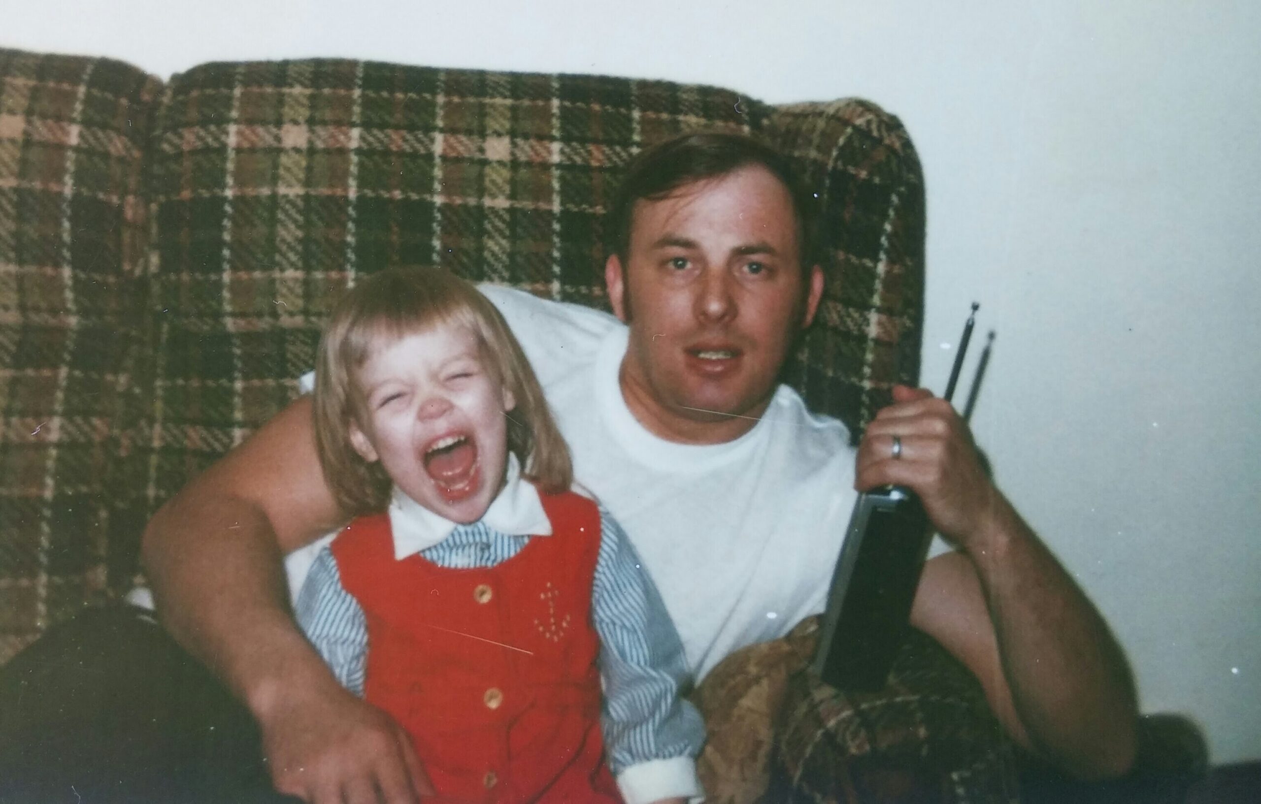 Father and young daughter laughing on a plaid couch; the man holds a bottle and a guitar neck.