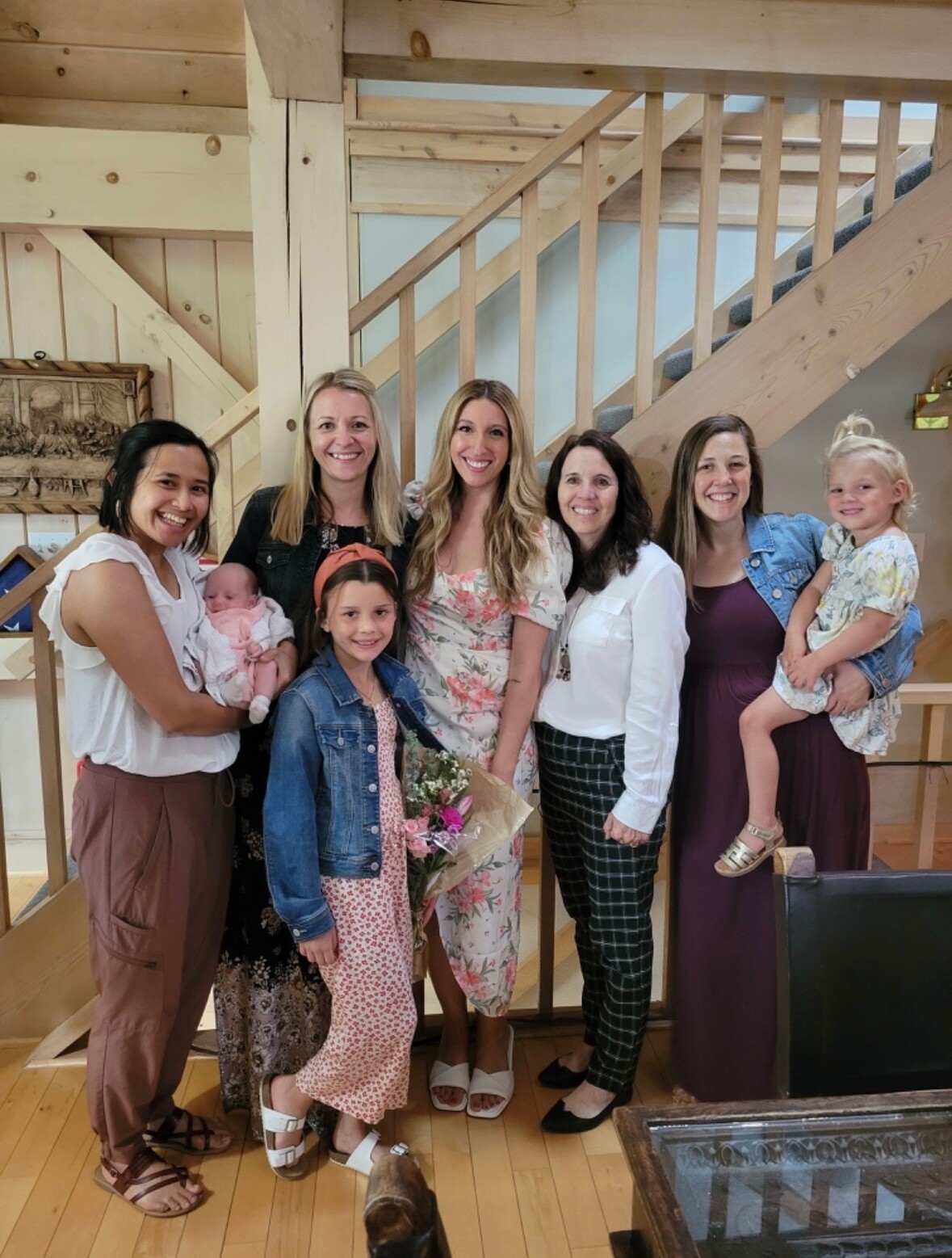 Group of nine people, seven adults and two children, smiling together indoors by a wooden staircase setting.