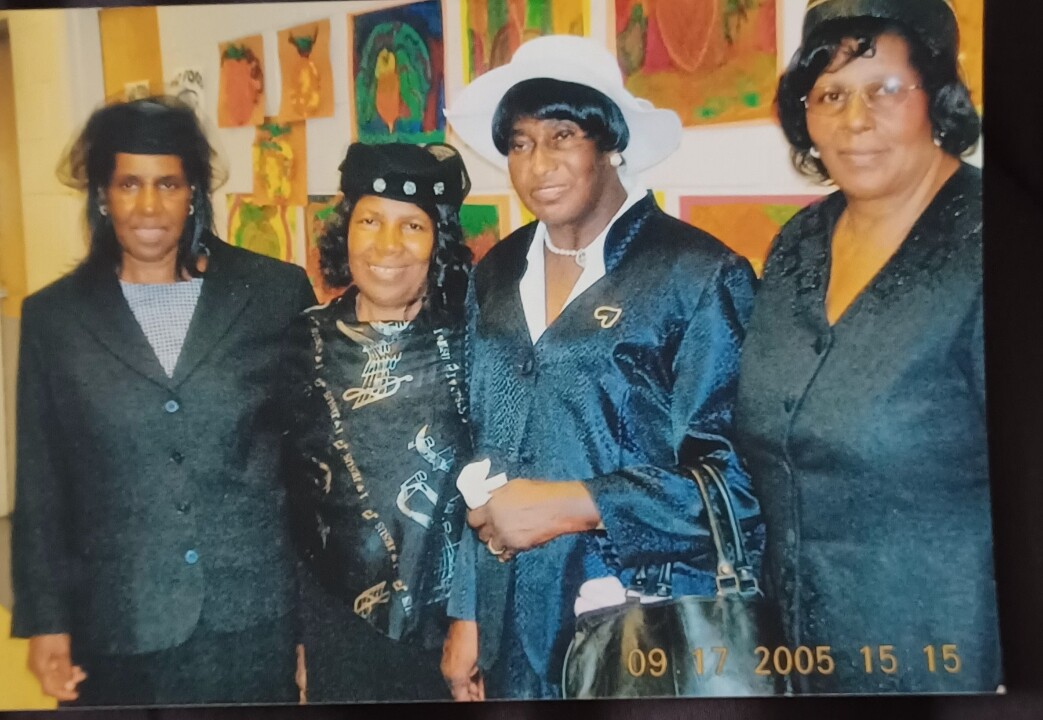 Group portrait of four adults in formal attire standing side by side at an event, colorful artwork on the wall behind them; date stamp reads 09/17/2005.