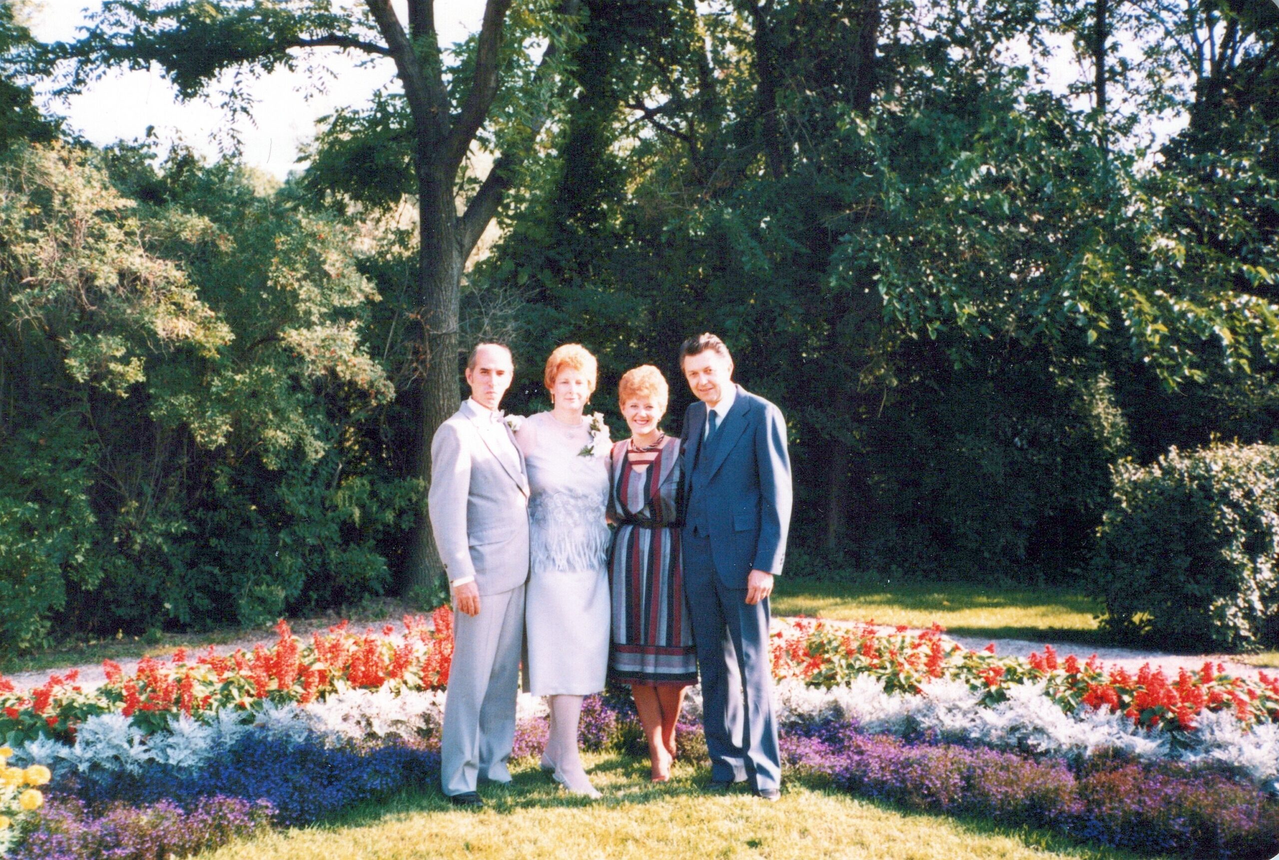 Four adults pose together in a garden with vibrant flower beds in front of green hedges and trees.