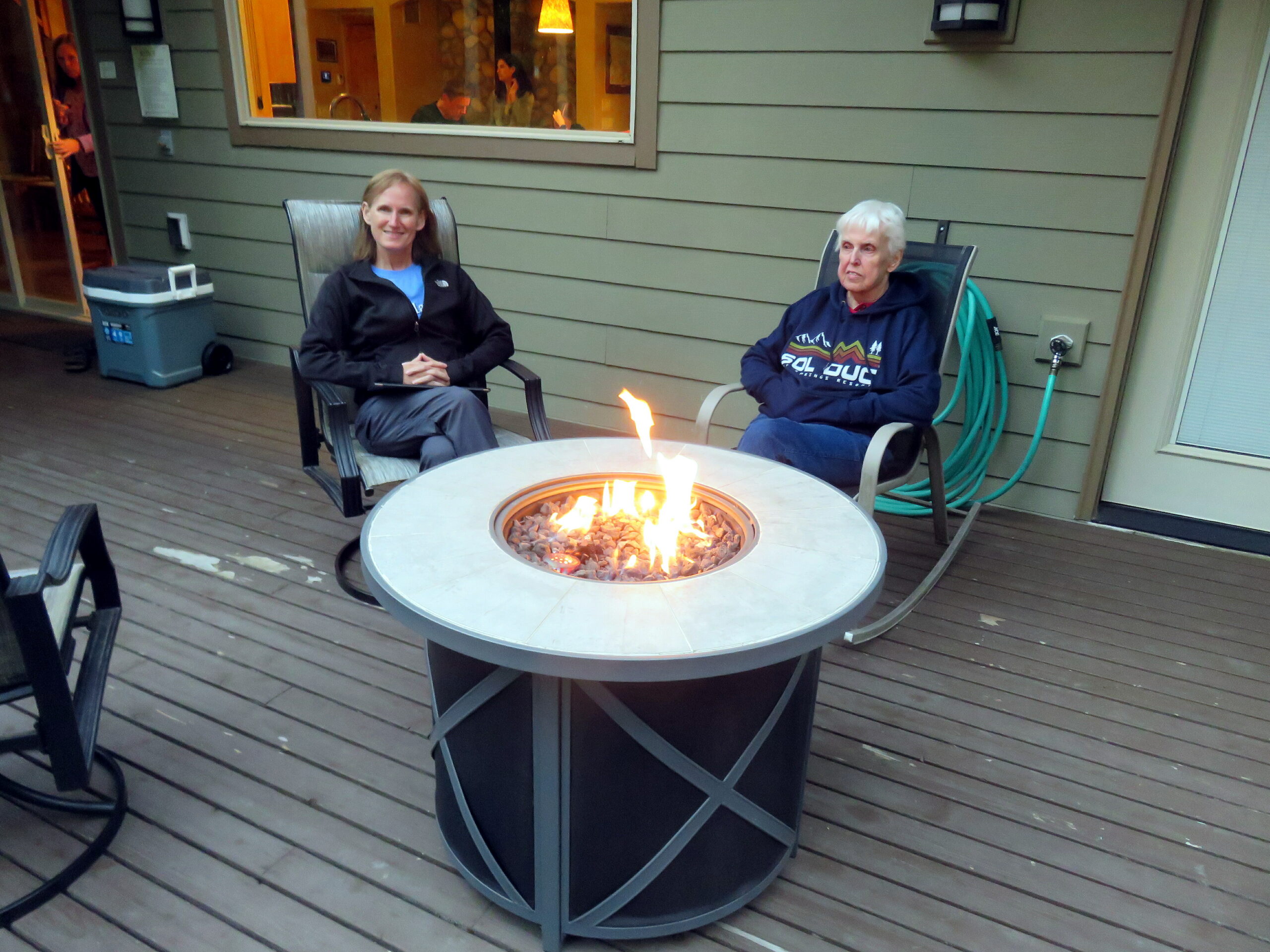 Two people sit in outdoor chairs around a glowing fire pit on a wooden deck outdoor patio.
