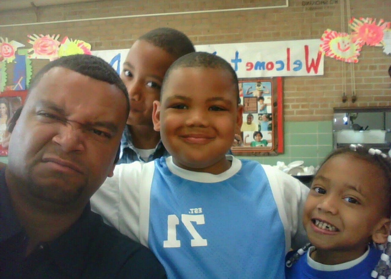 Three people posing for a selfie in a school cafeteria: an adult man on the left and two smiling kids in blue jerseys, with a colorful banner and wall photos behind them.