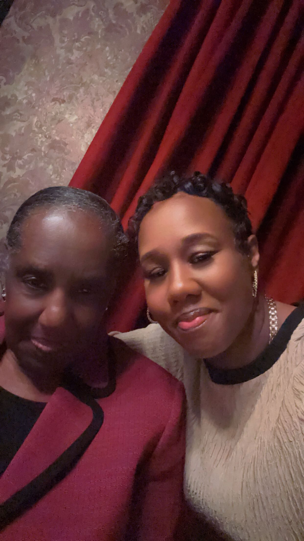 Two women posing closely for a photo indoors, burgundy curtains in the background.