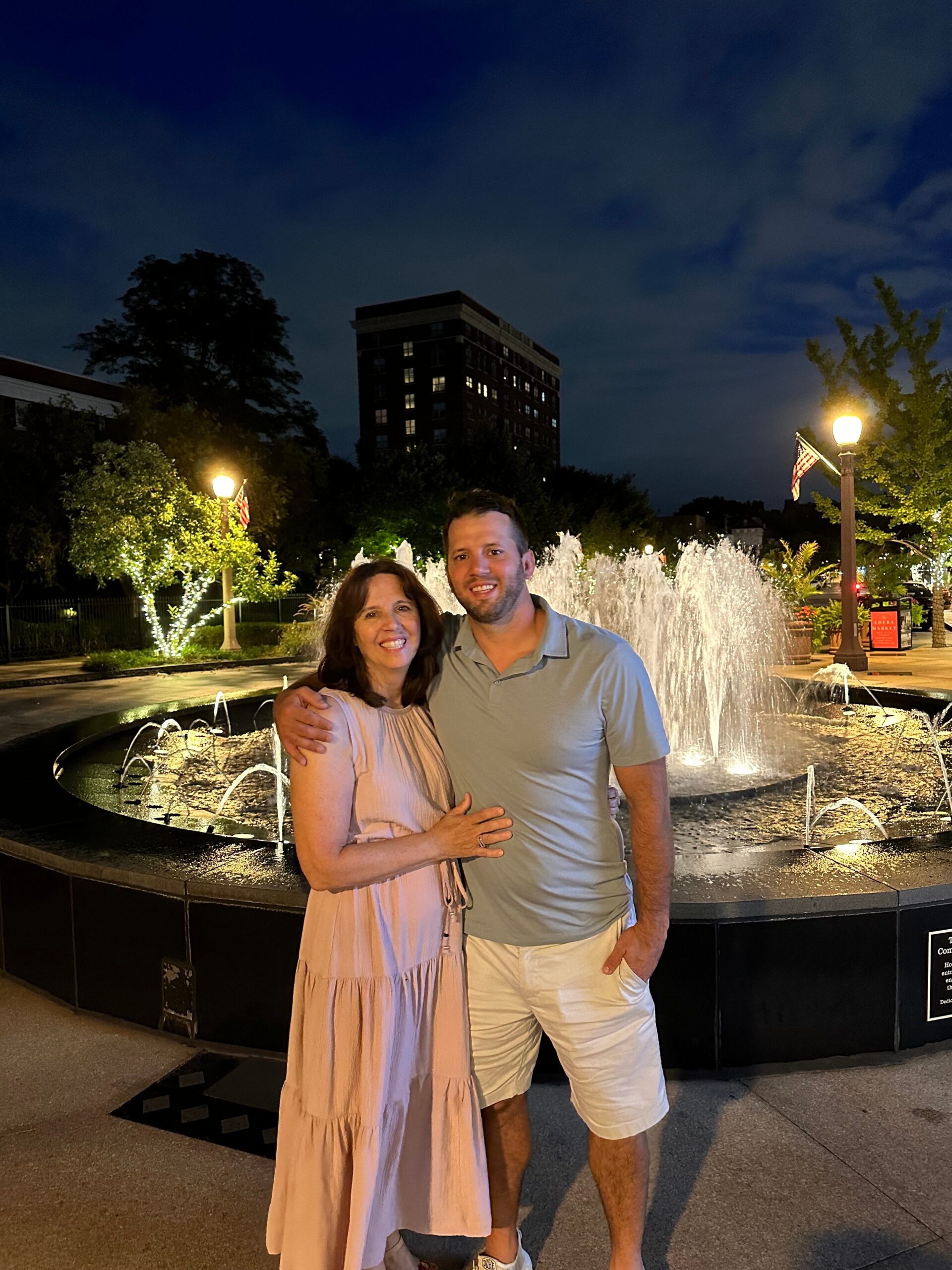 Couple standing arm in arm by a lit fountain at night in a city park. The woman wears a pink dress, the man a light blue polo and white shorts, both smiling.
