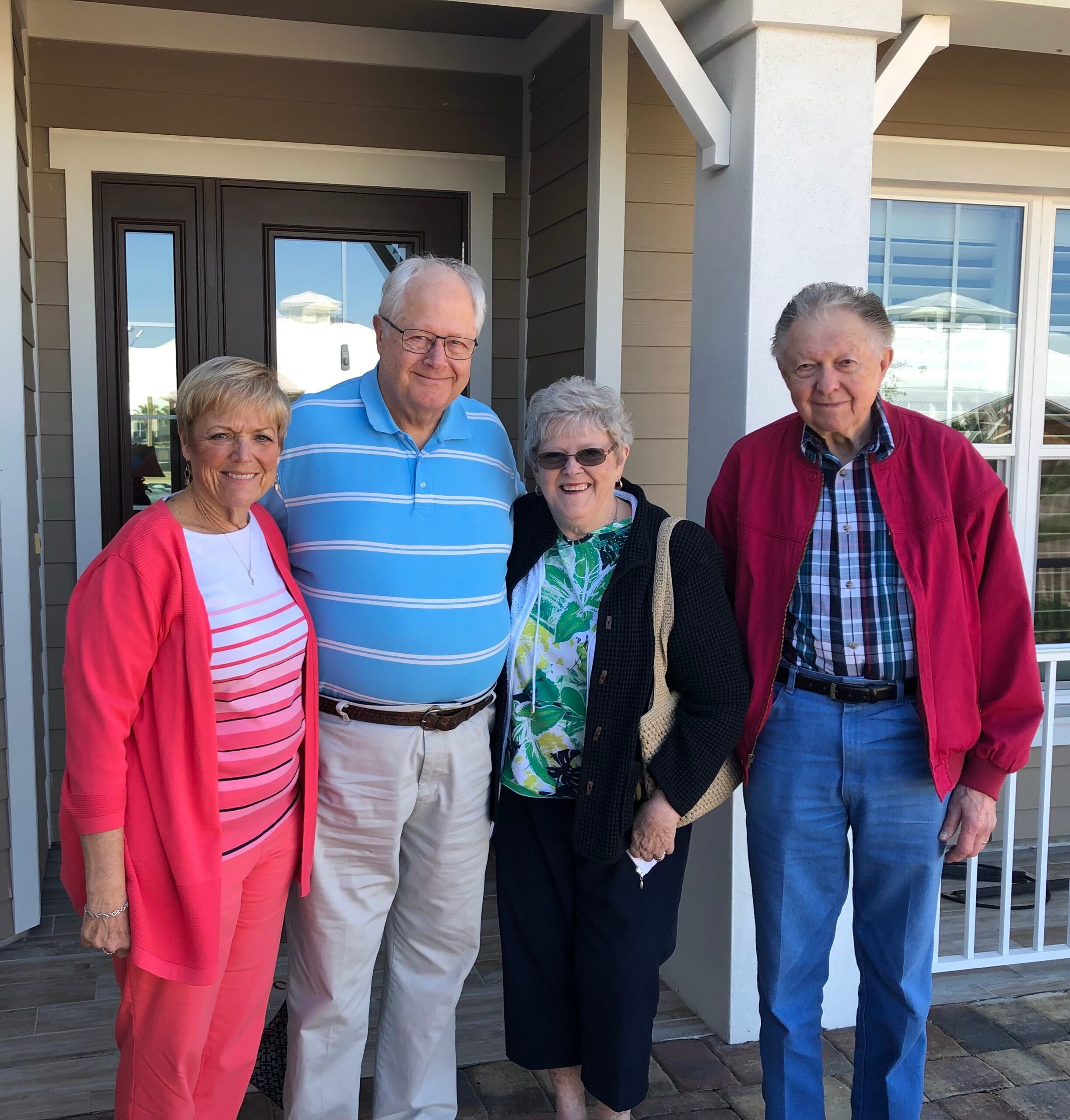 Smiling group of four older adults standing on a porch in front of a house, wearing casual colorful clothing (pink cardigan, blue striped polo, floral top, red jacket).