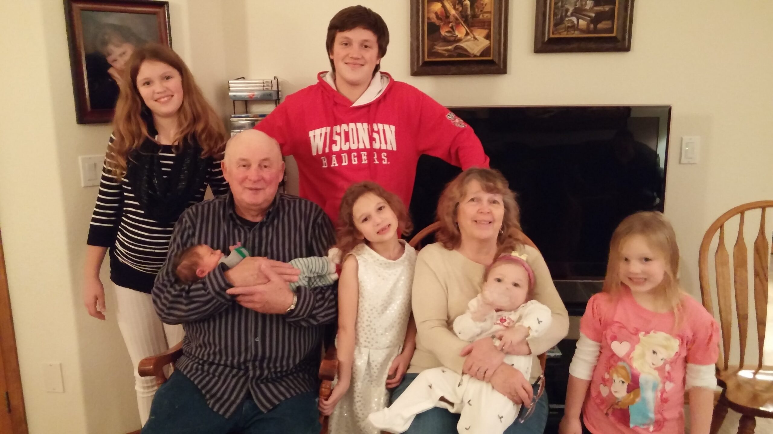 Family gathers for a group photo in a living room: an older man holds a newborn, adults and children stand or sit around him, a teenage boy in a red Wisconsin hoodie in back, framed art on the wall.