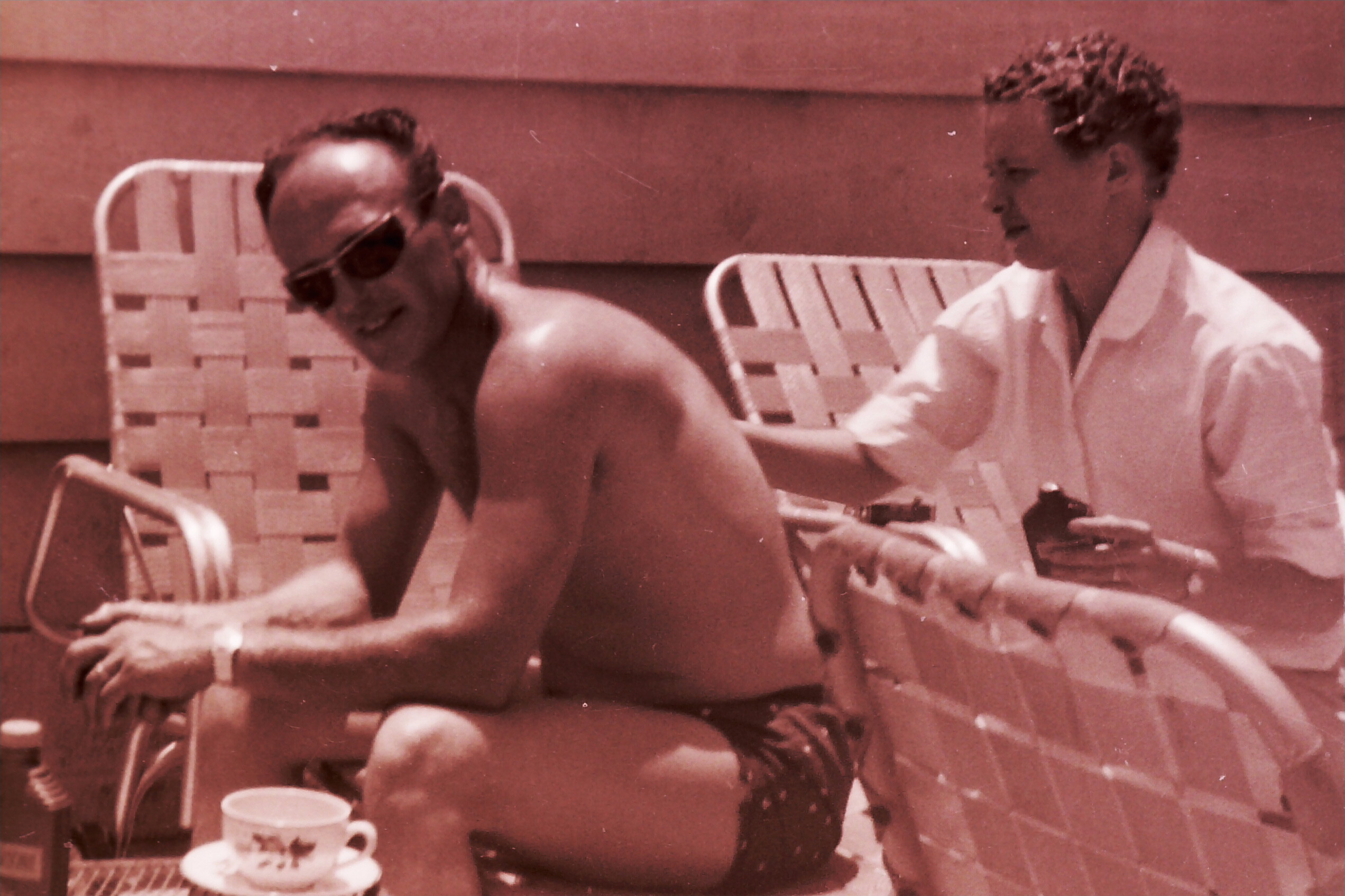 Two men at a backyard gathering by the pool: one shirtless with sunglasses, the other in a white shirt, seated on lawn chairs and talking.