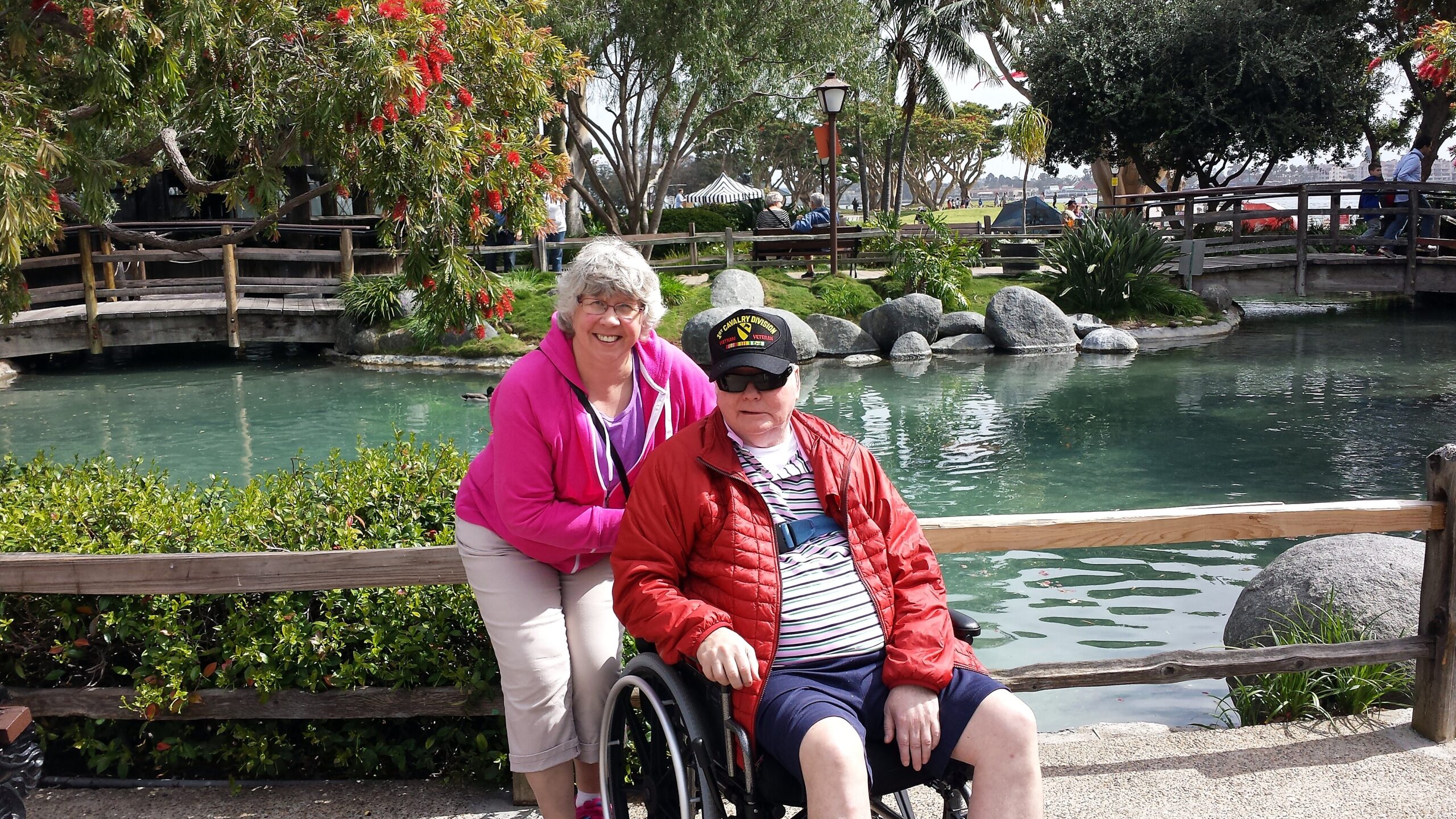 Two elderly people by a pond in a park: a smiling woman in a pink jacket stands behind a man in a wheelchair, with a wooden fence and greenery around them.