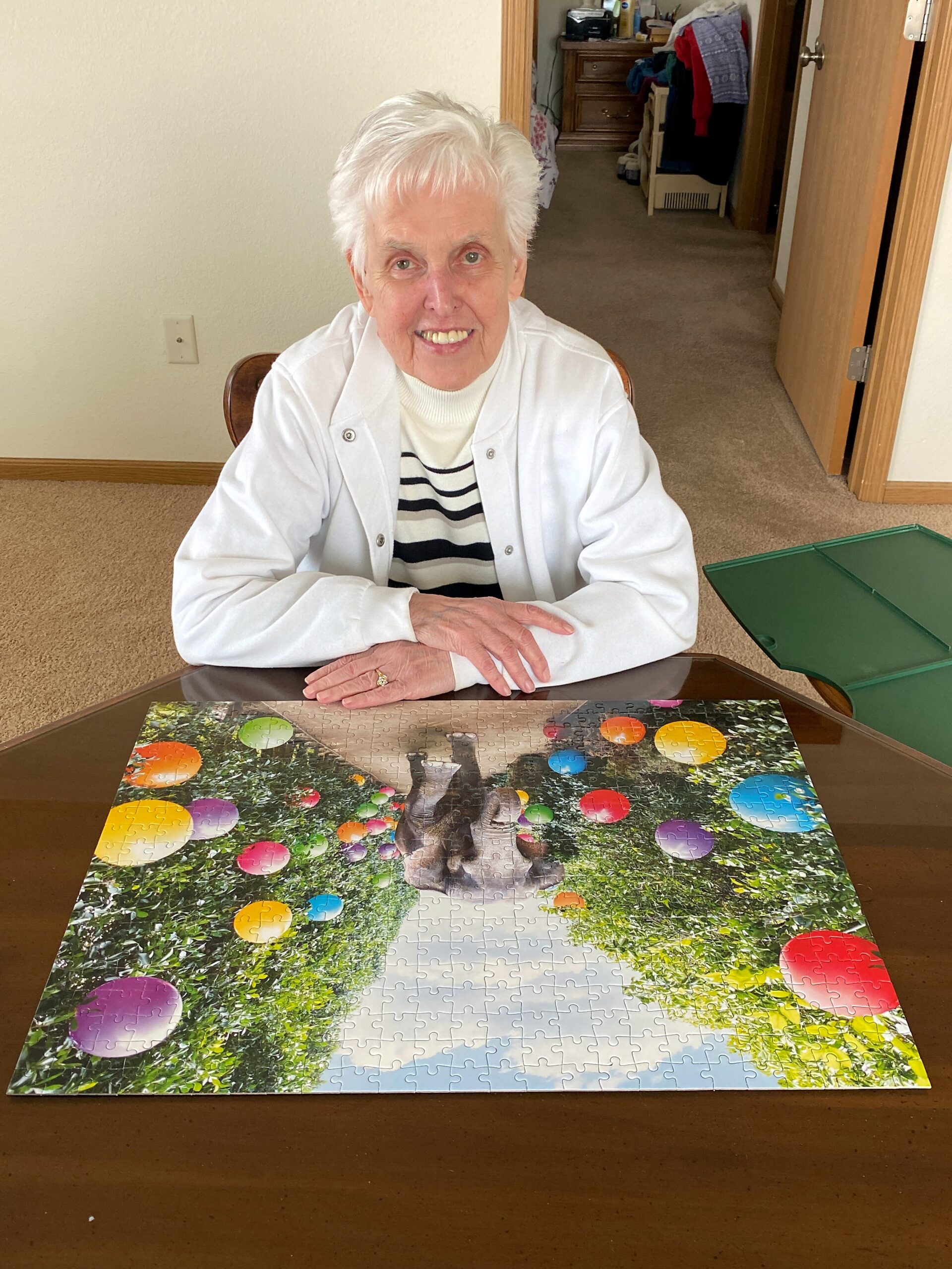 Older woman with white hair smiling, seated at a table with a colorful jigsaw puzzle spread out in front of her.